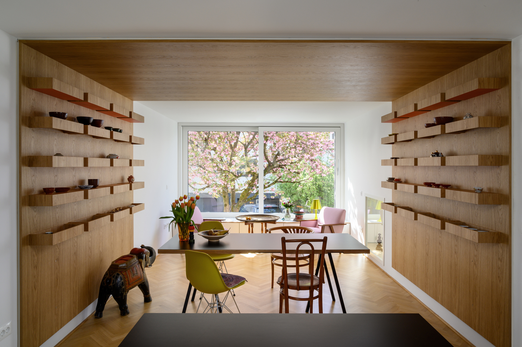 Dining room framed by oak shelving walls and herringbone floor, with a blossoming pink tree visible through the sliding window