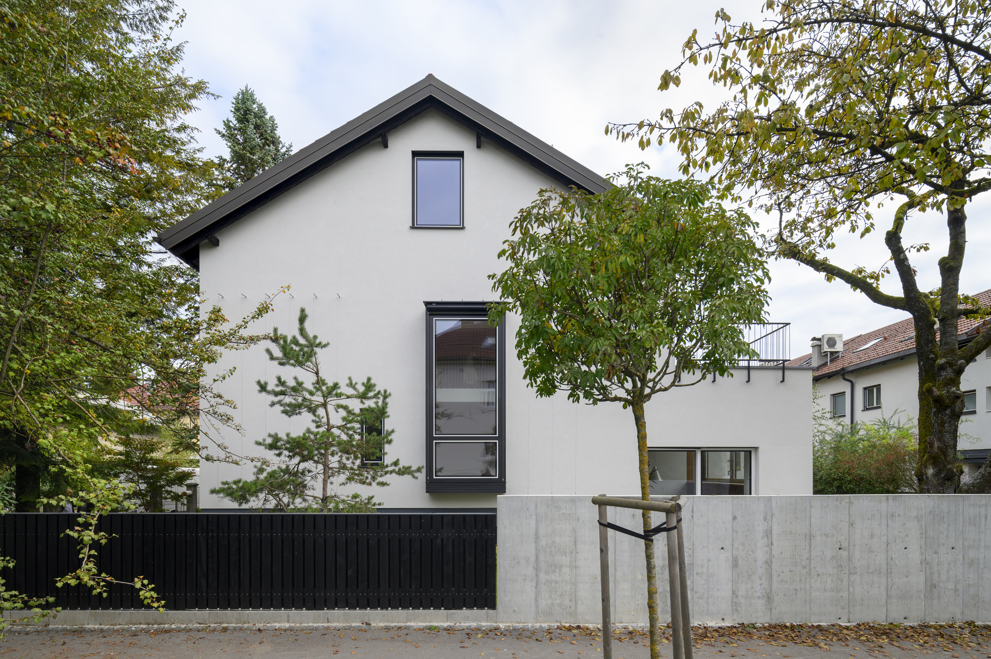 Gabled white side facade with a tall black-framed window, a black timber fence and concrete wall, seen among autumn leaves