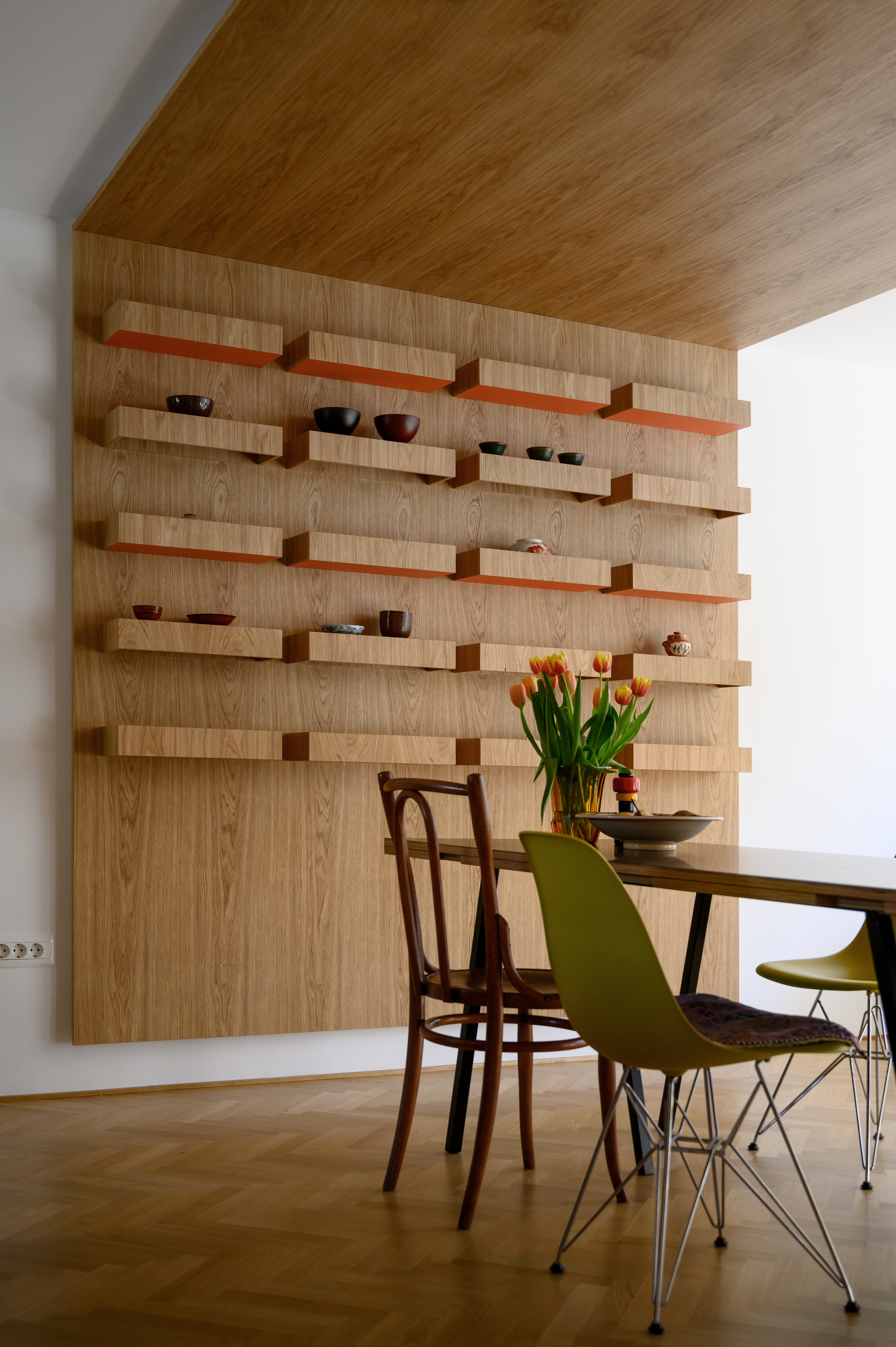 Oak veneer wall with staggered floating shelves painted orange underneath, holding ceramic bowls above a dining table with tulips in soft daylight