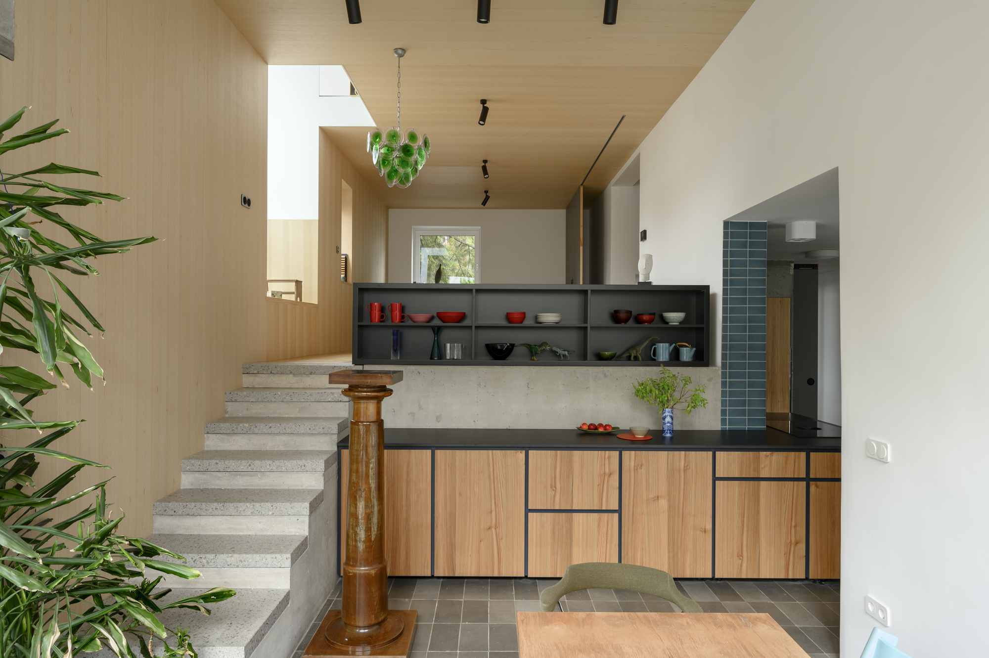 Terrazzo steps rising beside a kitchen with oak cabinet fronts, a dark open shelf and a green glass pendant, with a leafy plant in the foreground