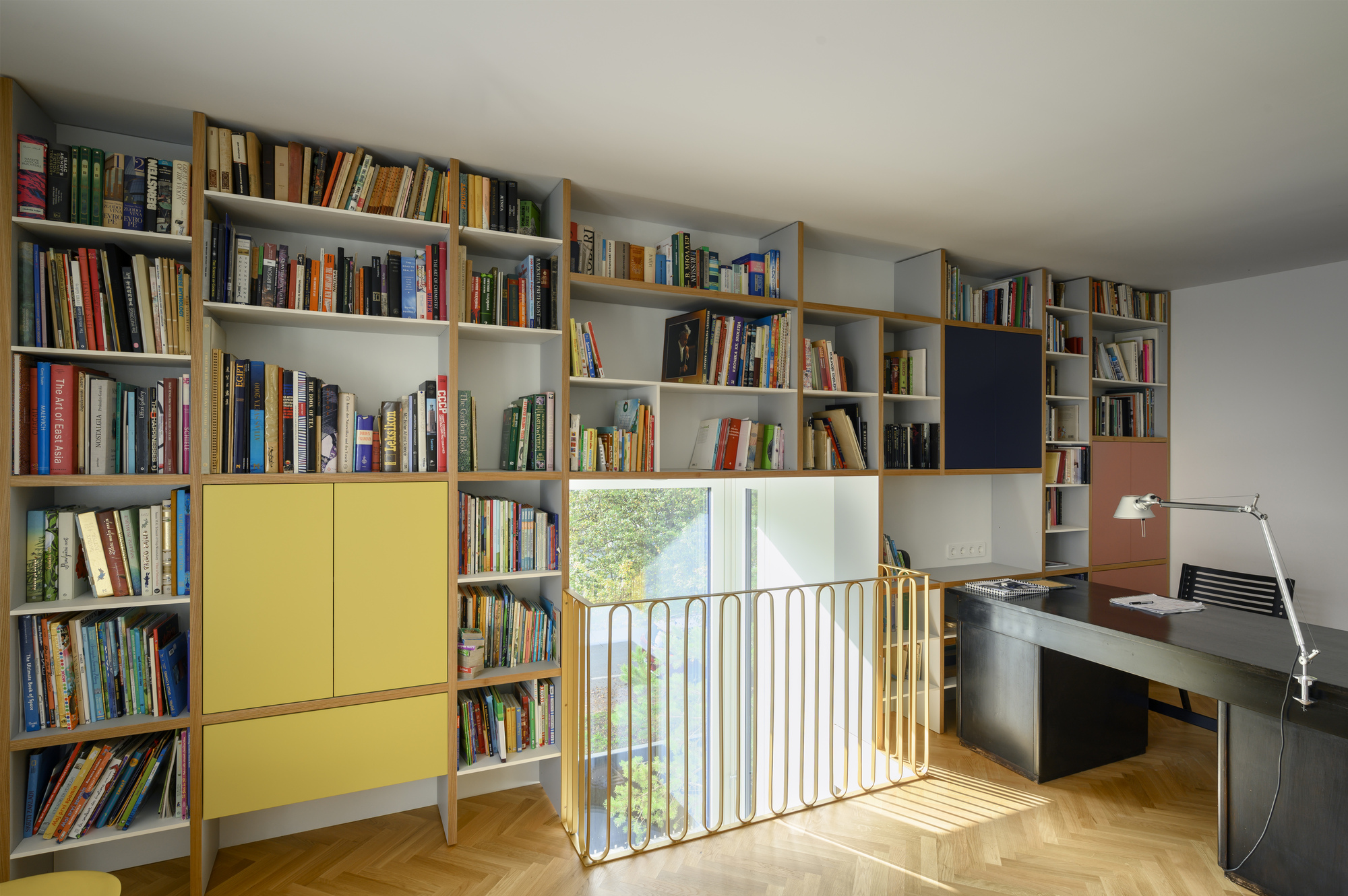 Wall of crowded bookshelves with yellow cabinet doors beside a brass-toned balustrade, with afternoon sun striking the herringbone floor