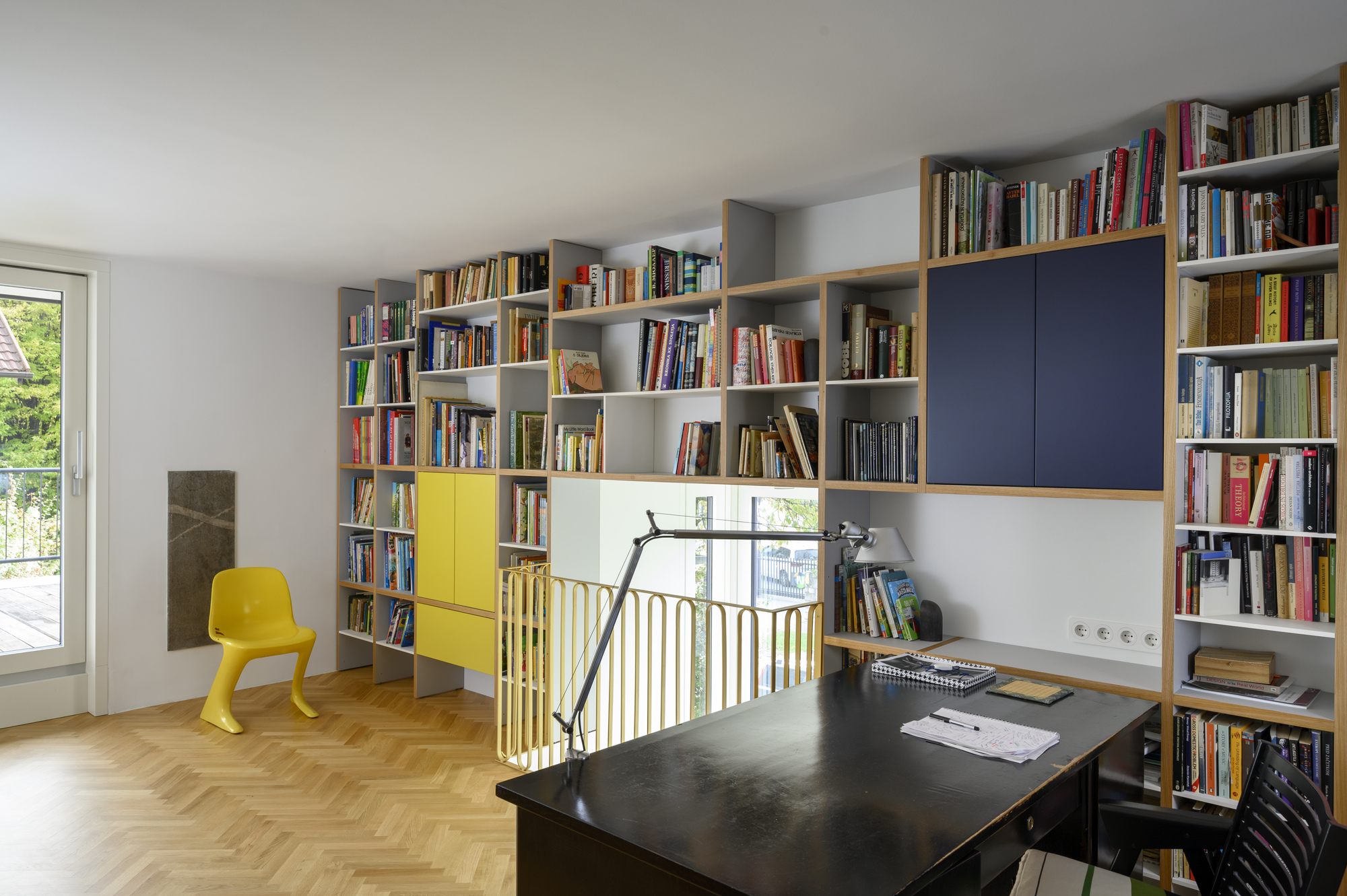 Study with floor-to-ceiling shelving, navy and yellow cabinet doors, a black desk with a task lamp and a yellow chair by the balcony door