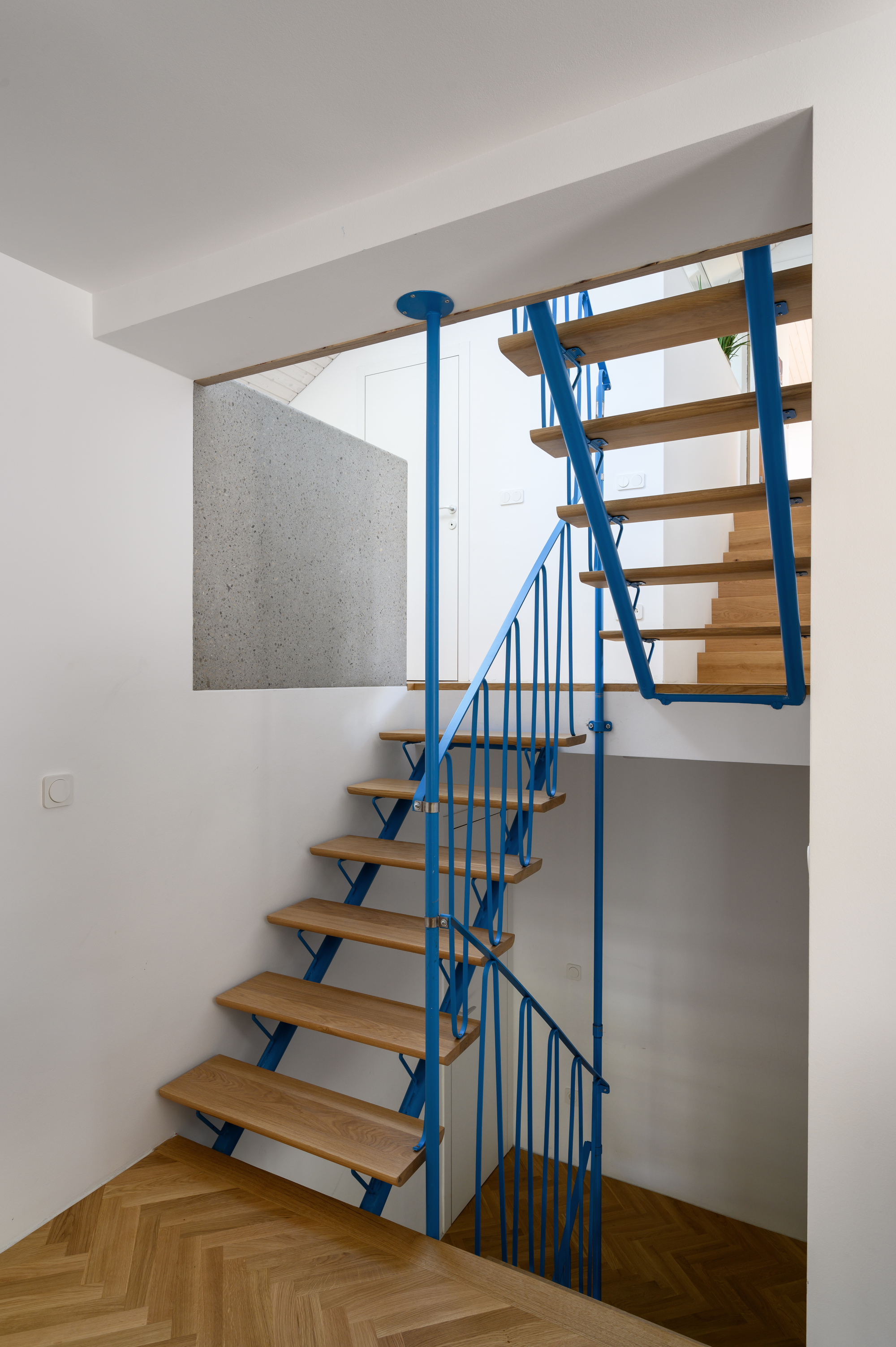 Open-tread staircase with oak steps and bright blue steel railings turning past a rough terrazzo wall, above a herringbone parquet landing