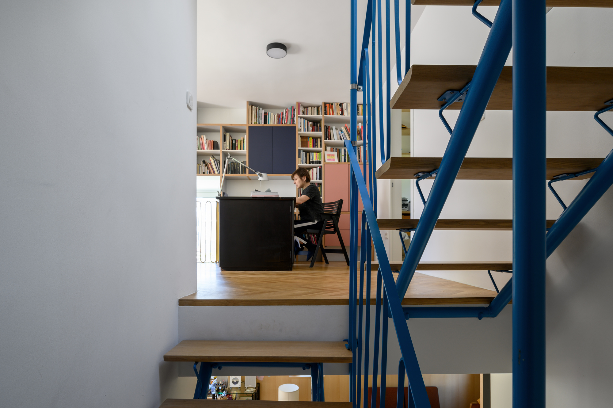 View through a blue steel stair with timber treads to a person working at a black desk before bookshelves