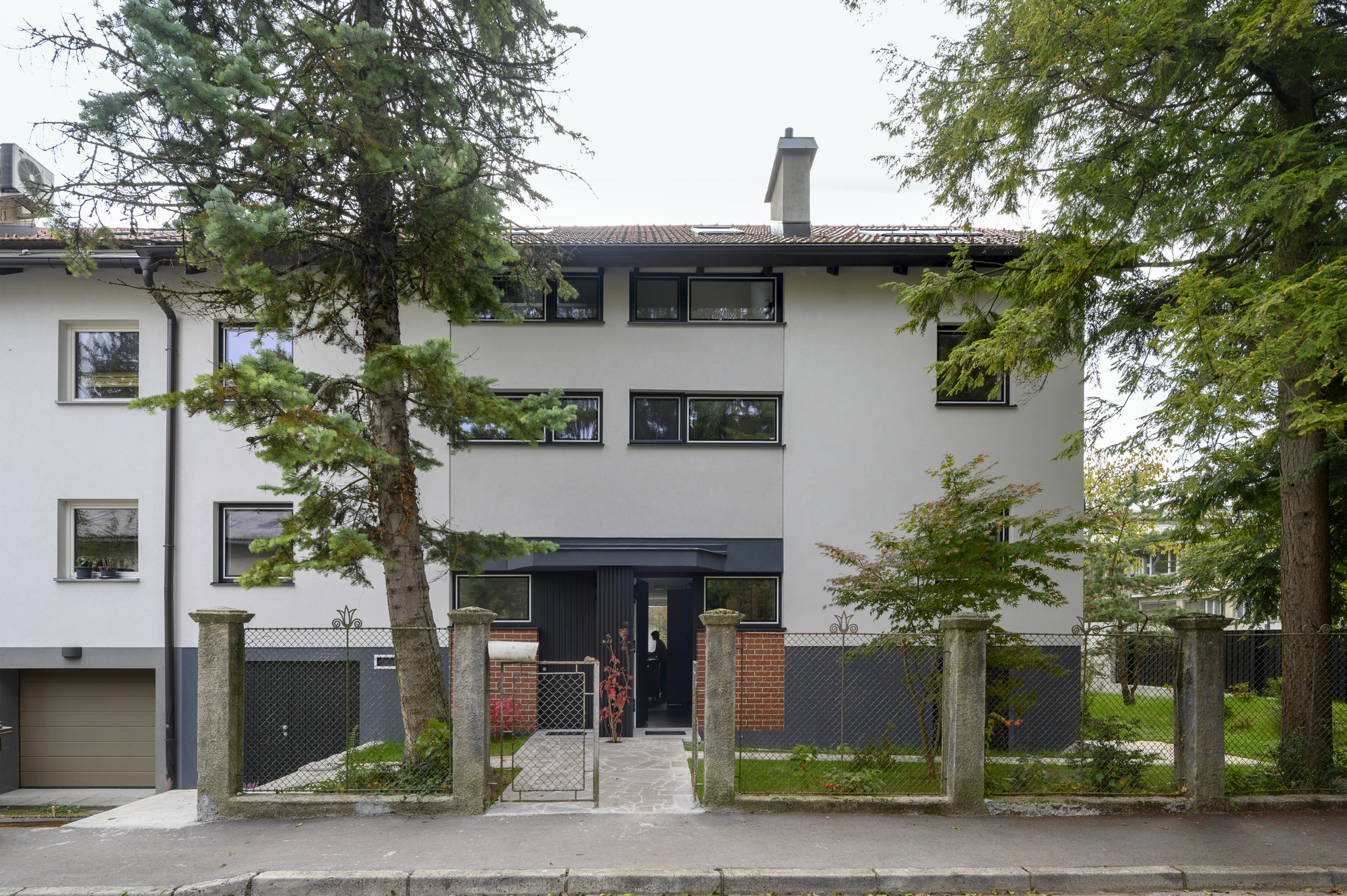 Street facade of the three-storey rendered house behind a wire fence and brick-and-concrete gate posts, framed by conifers in overcast light