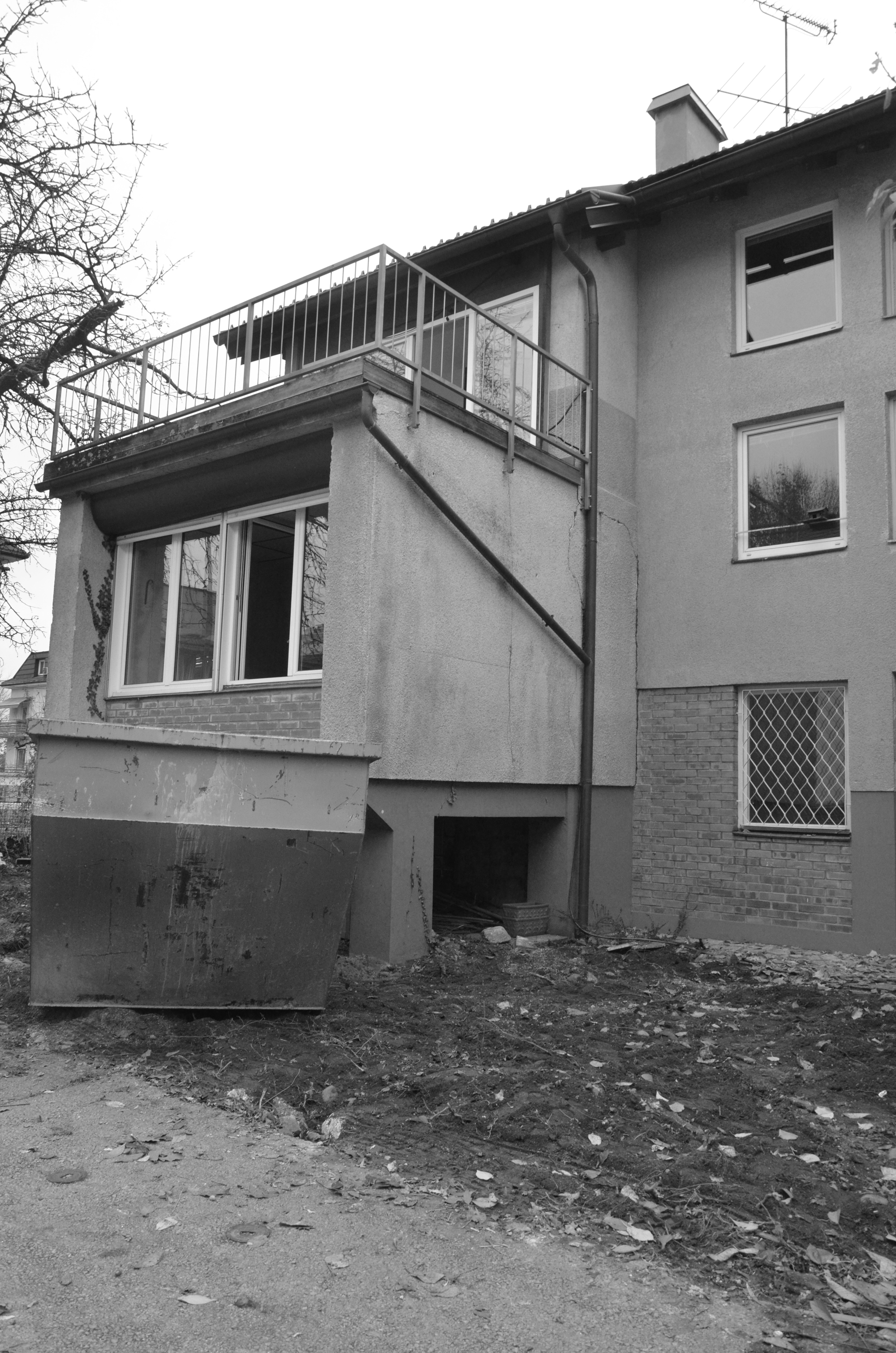 Black and white rear of the original house with a balcony over a bay window, a skip bin and bare ground in winter