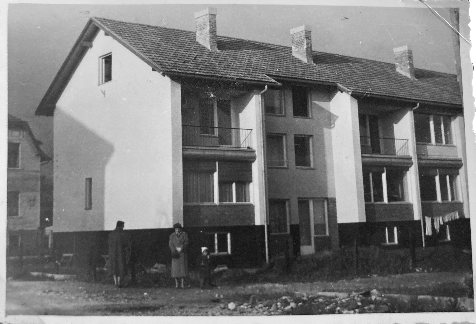 Archival photograph of the terraced housing block with tiled roof and chimneys, three people standing on rough ground in front