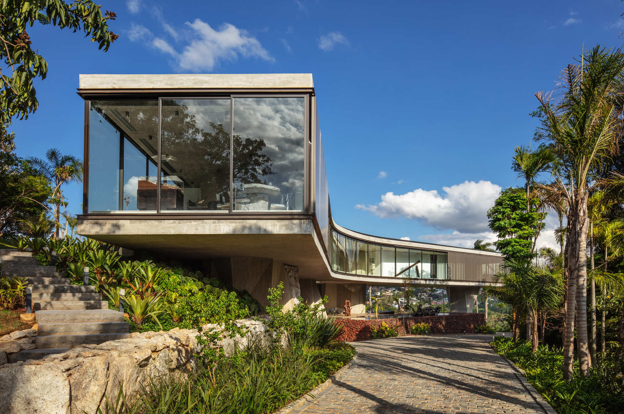 Cantilevered concrete box with floor-to-ceiling glazing above a stone-edged garden bed and cobbled drive, under a blue sky with palms