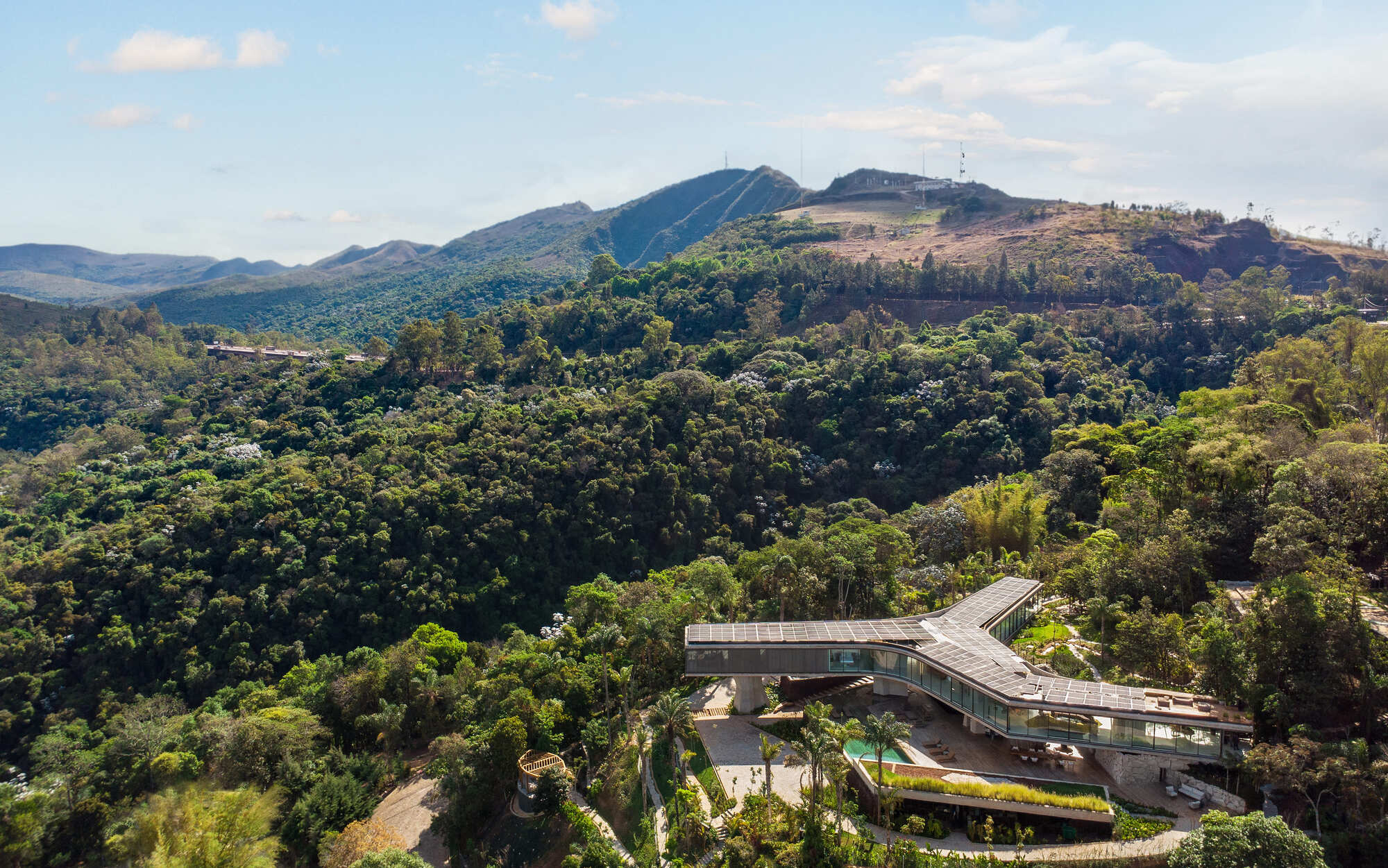 Aerial view of the solar-panel roof and glass bar emerging from dense forest, with hazy mountains behind in daylight