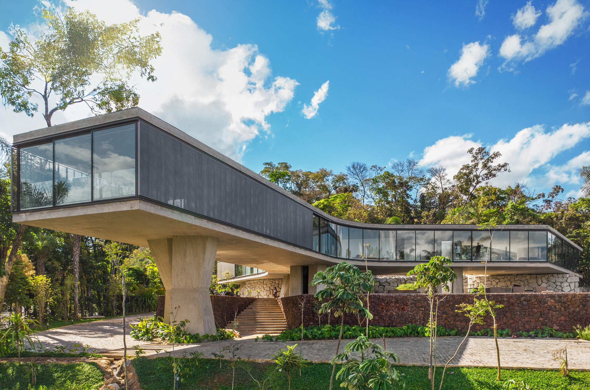 Long concrete volume on angled piers with a dark cladded flank and curving glass, set above a red stone retaining wall