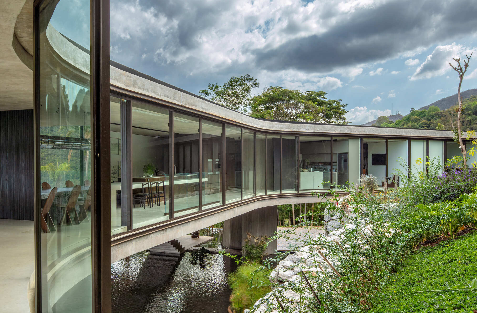 Curved glazed corridor with slim bronze mullions bridging over a dark reflecting pond, with stormy clouds and green hillside beyond