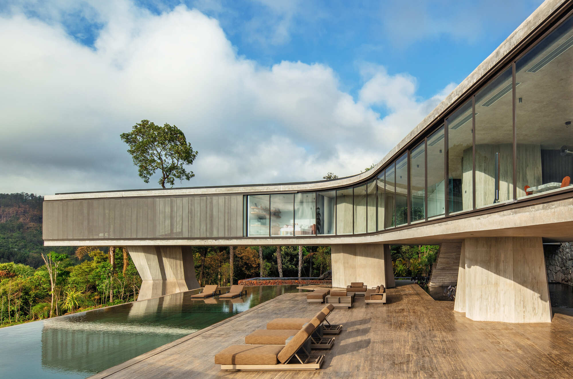 Timber pool deck with lounge chairs beneath the raised concrete bar, its tapered piers reflected in the infinity-edge water