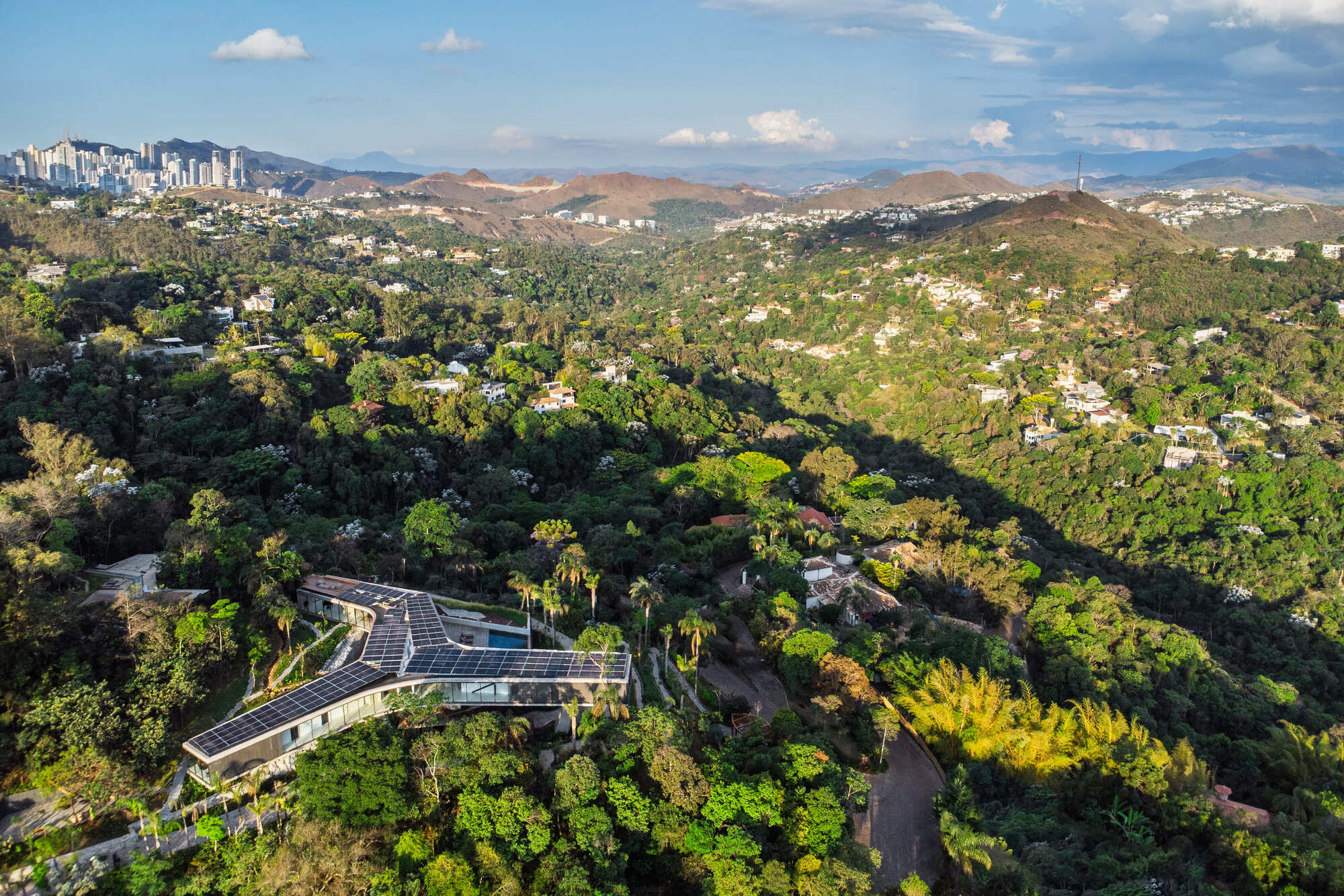 High aerial view of the solar-roofed house in a forested valley, with a distant city skyline in late afternoon light