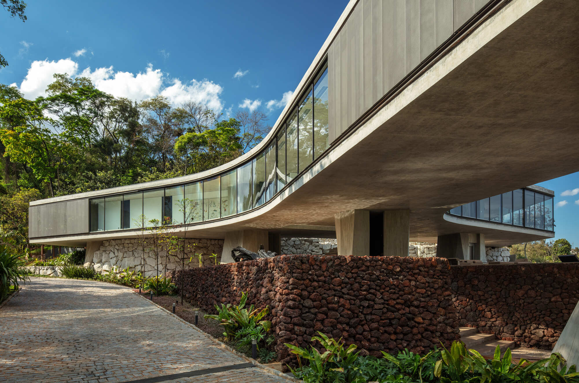 Curving concrete volume with a glazed ribbon window cantilevered over a red volcanic stone wall and cobbled drive, under a blue sky with scattered clouds