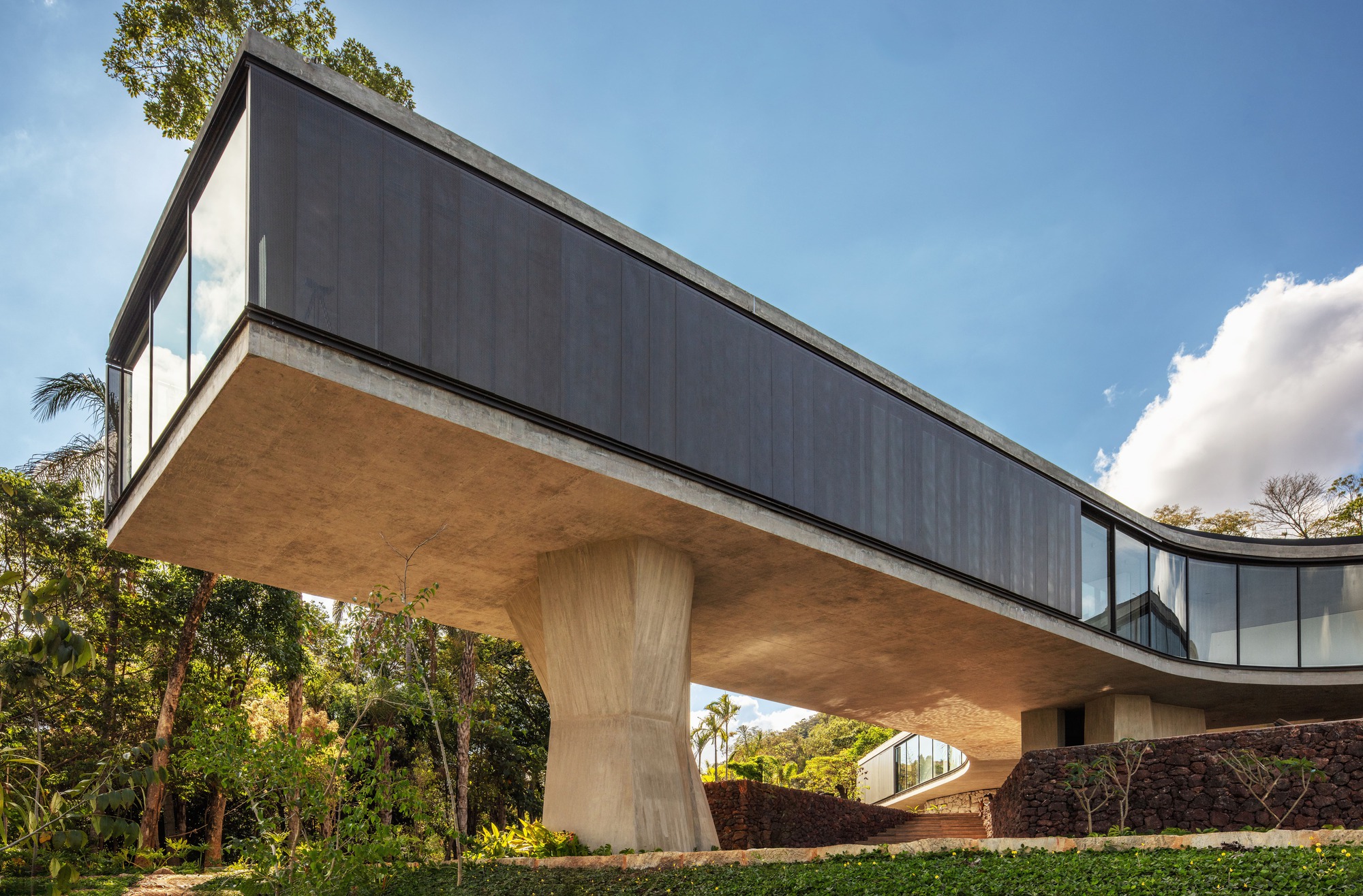 Low-angle view of the cantilevered concrete box with dark cladding resting on a faceted column, framed by forest and bright clouds