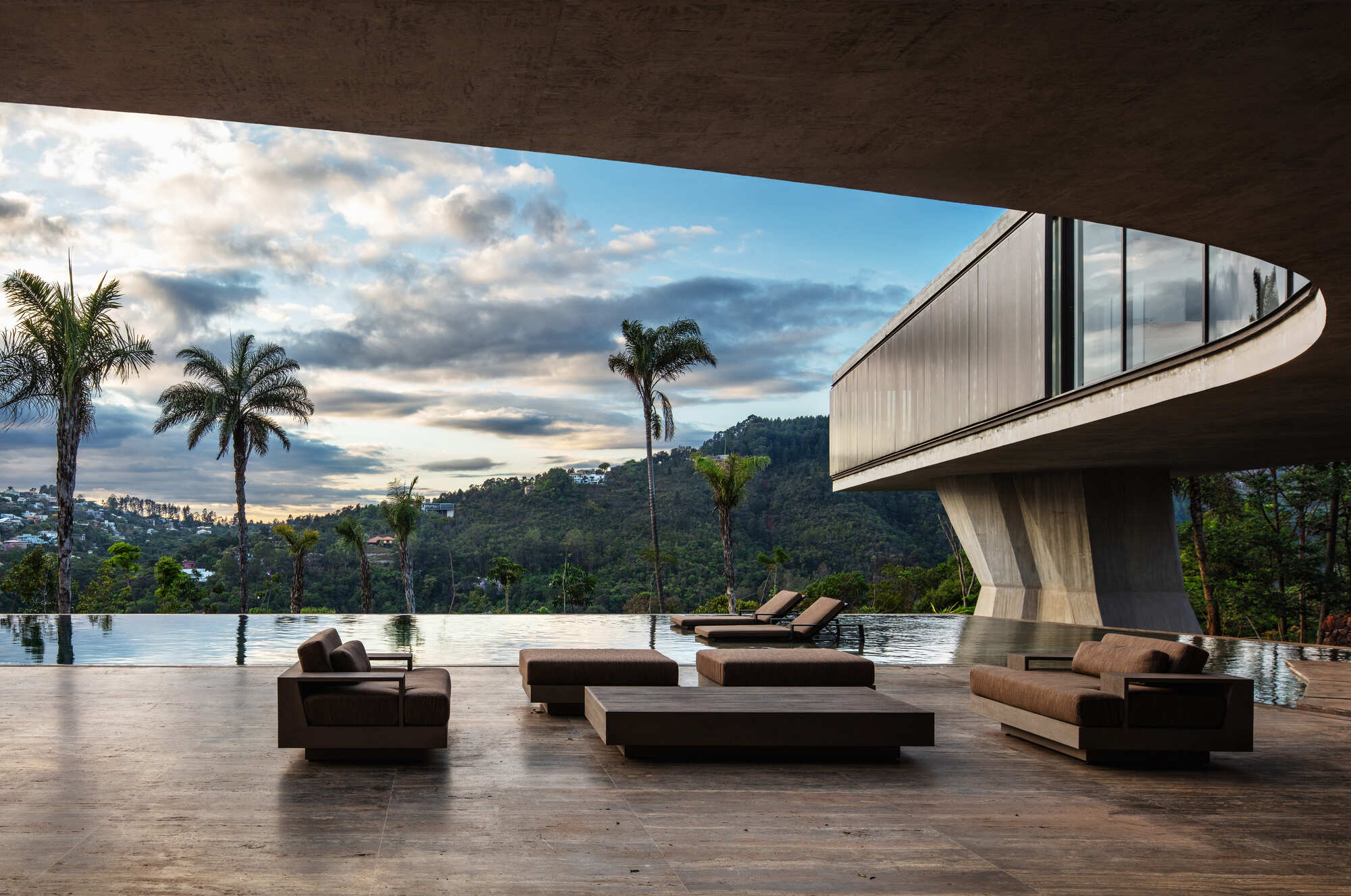 Covered terrace with brown lounge sofas facing an infinity pool and palm trees, with a cloudy dusk sky over forested hills
