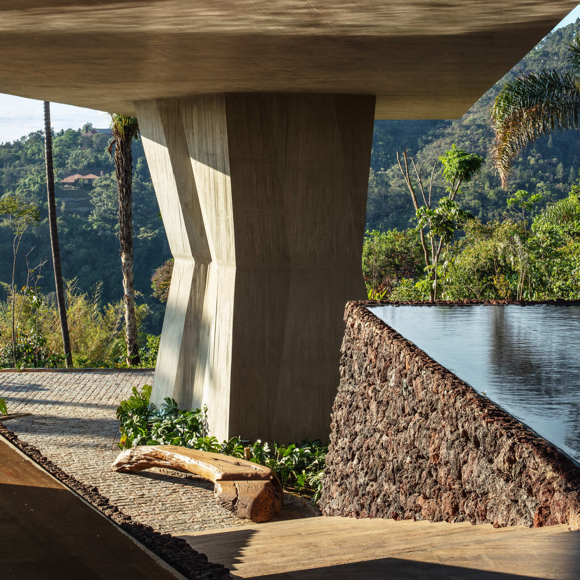 Faceted concrete column beside a red volcanic stone pool wall, with a driftwood bench on cobbles in raking sunlight