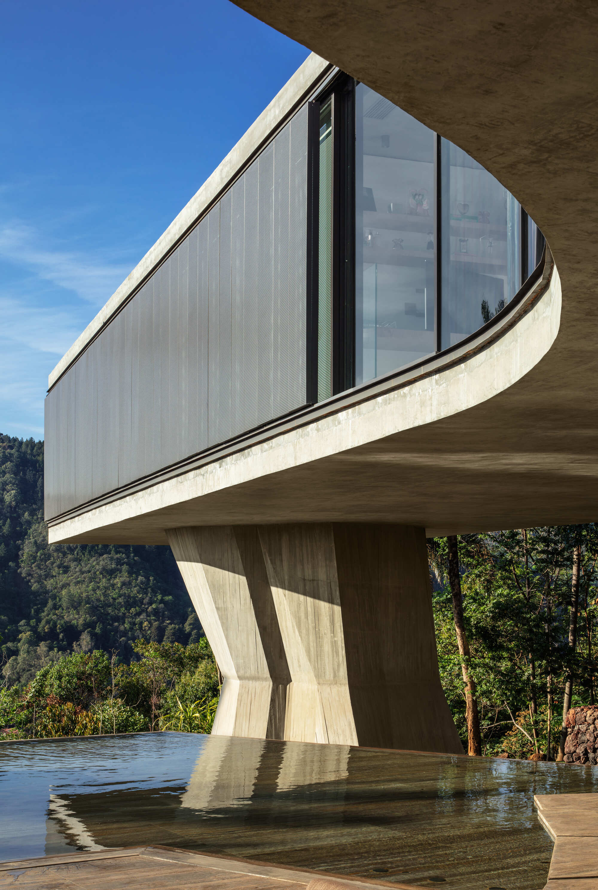 Curved concrete slab edge and dark clad volume above a faceted column reflected in still pool water under clear blue sky