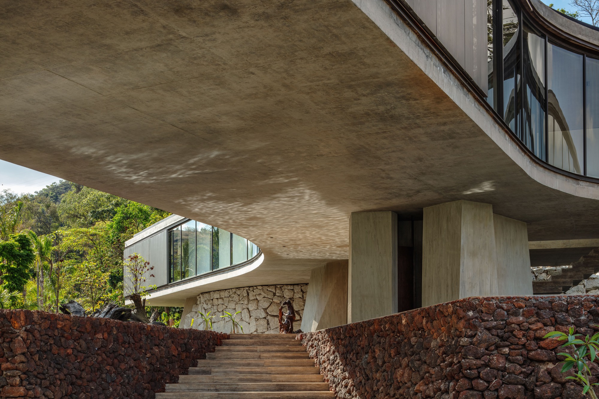 Timber steps rising between red volcanic stone walls beneath a concrete soffit dappled with reflected light, with forest beyond