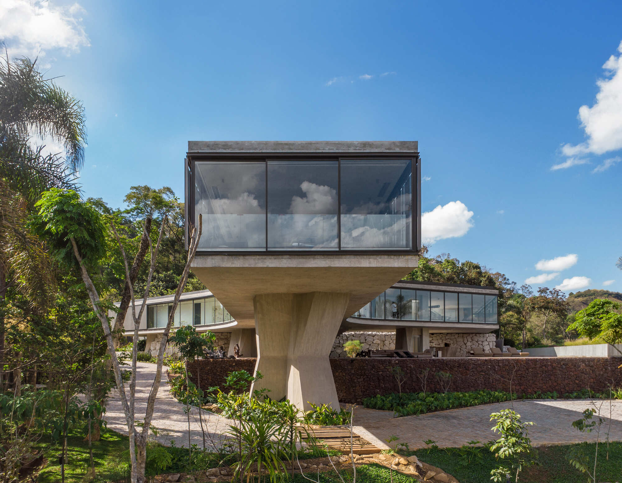 Glass-walled box cantilevered on a faceted concrete column above a stone-paved path, with low curved wings behind under a blue sky with clouds