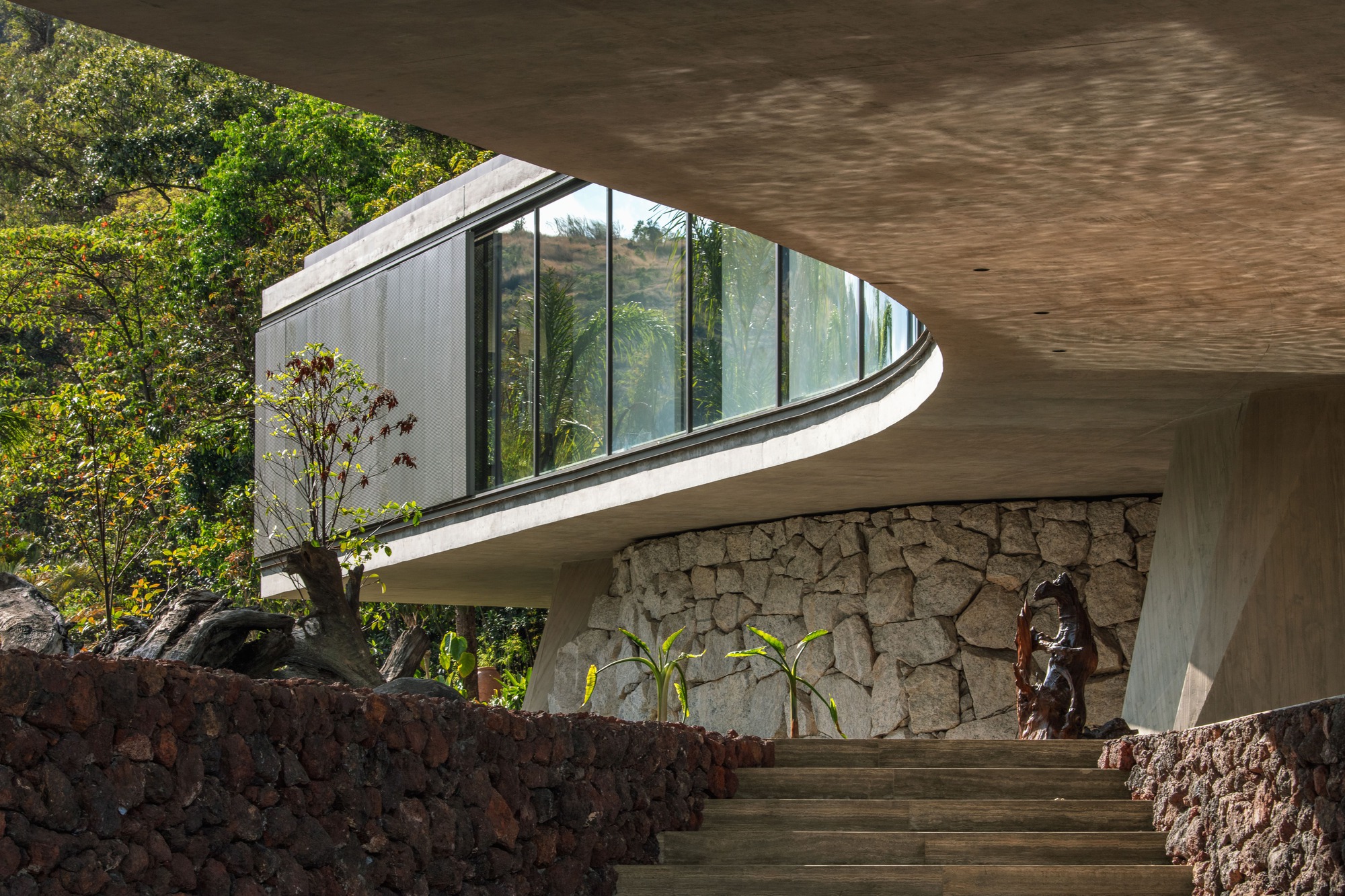 Curved concrete slab and glazed volume above a rubble stone wall, with stone steps and a carved wood sculpture in dappled light