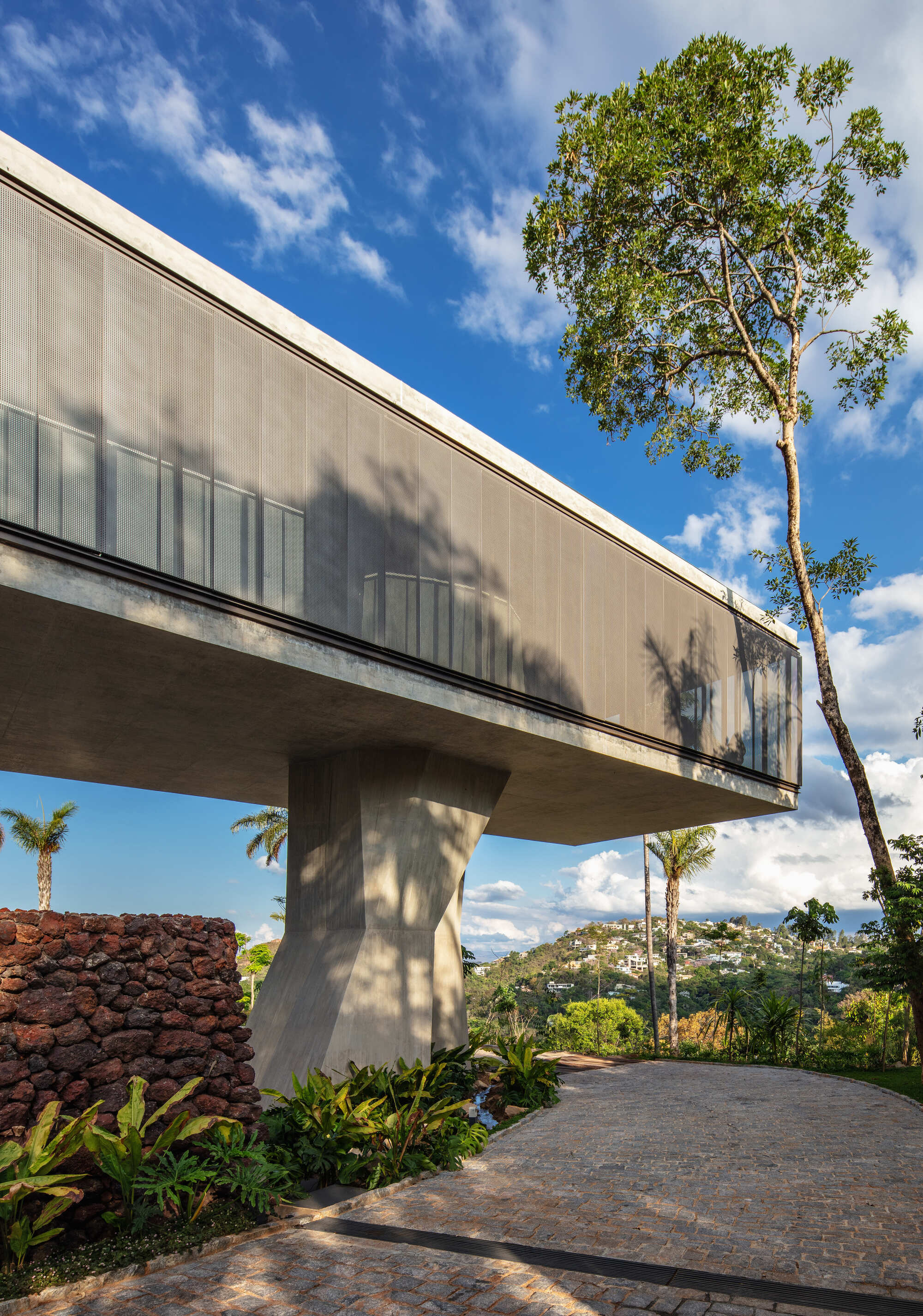 Side view of the cantilevered concrete volume with a screened facade, resting on a faceted column beside a red volcanic stone wall