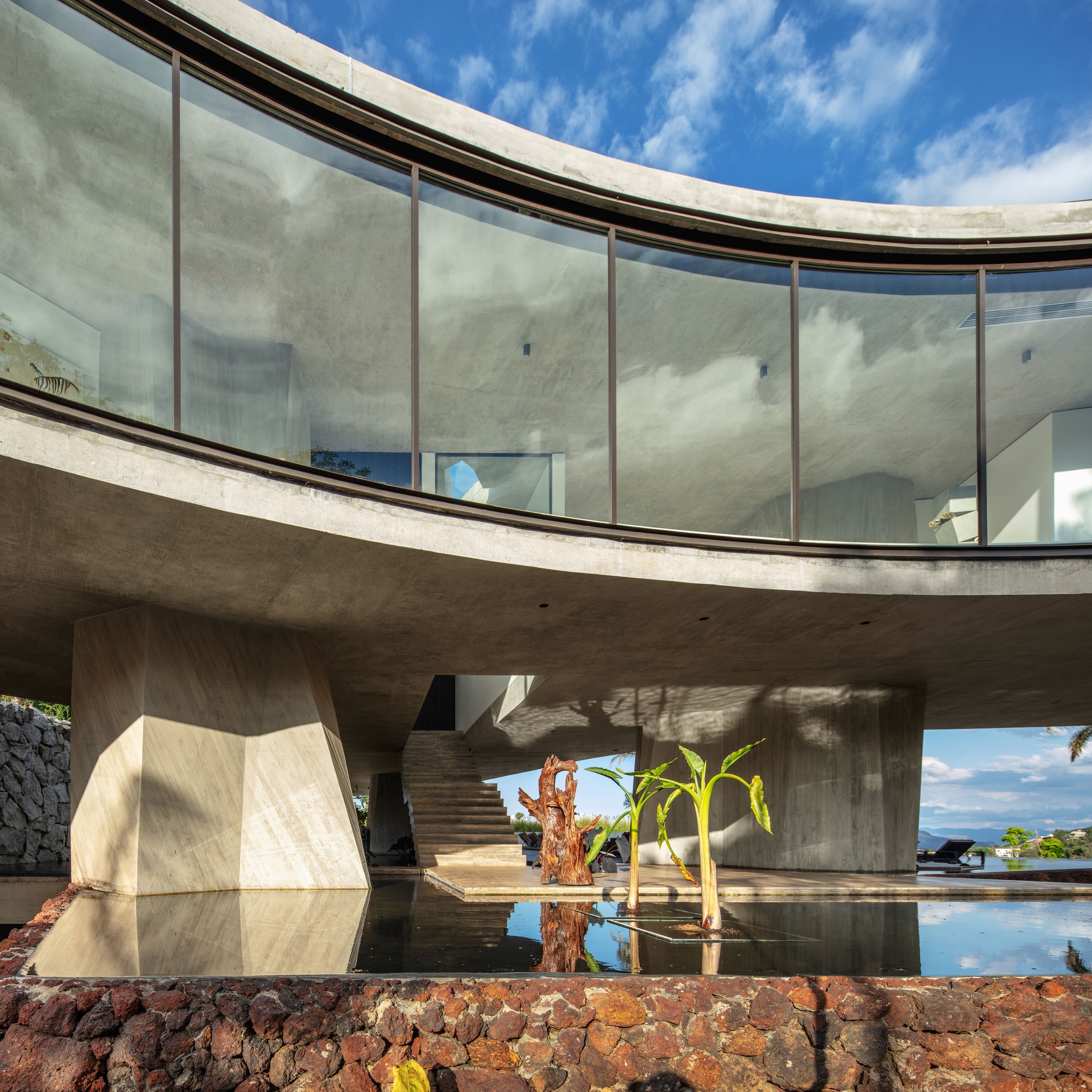 Curved glazed band above faceted concrete columns and a reflecting pool with banana plants, a driftwood sculpture and a floating stair behind