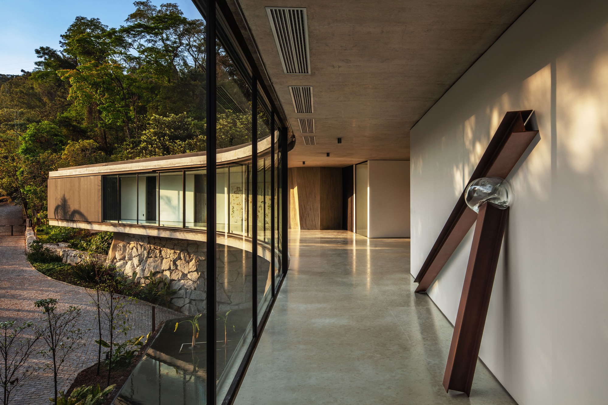 Interior gallery along a curved glass wall with polished concrete floor and a leaning rusted steel beam sculpture against a white wall
