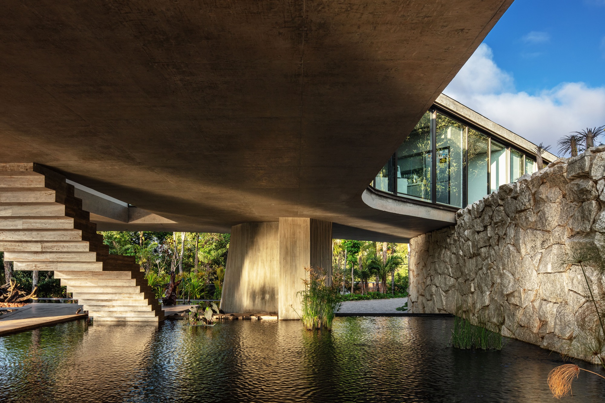 Underside of the concrete slab above a still pond, with a cantilevered stair, a round column and a rubble stone wall in afternoon light