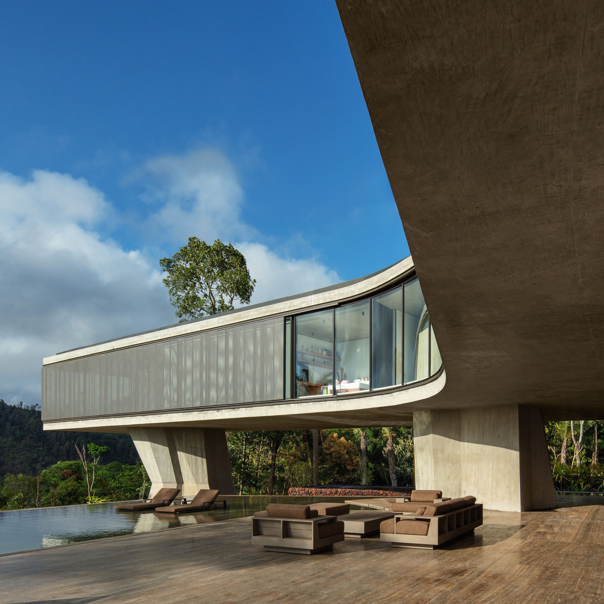 Curved concrete volume raised on angular piers above a terrace with lounge seating and an infinity pool under blue sky