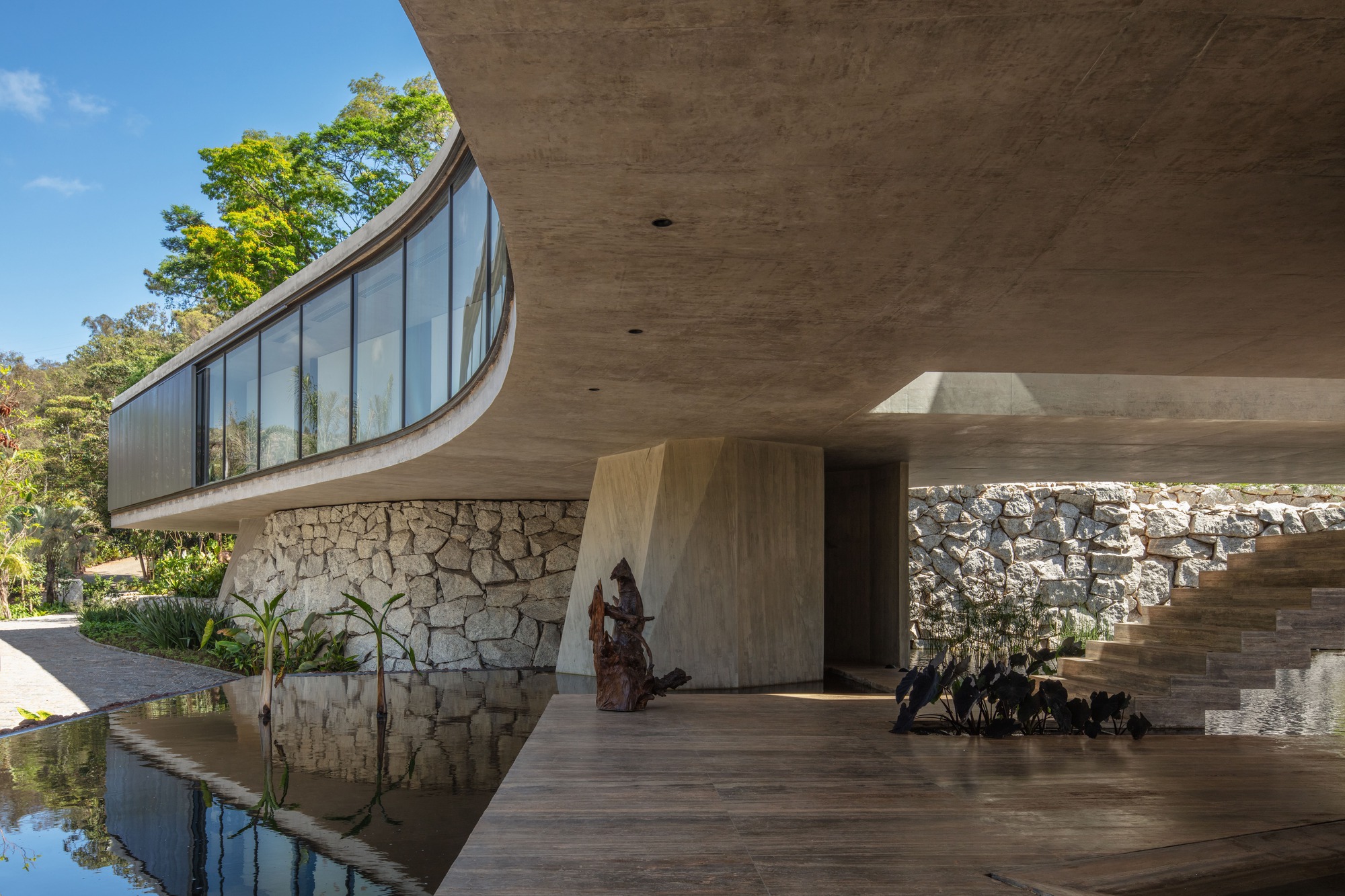 Curving glazed upper volume over a rubble stone wall and shallow water channel, with a sculpture standing on the timber deck