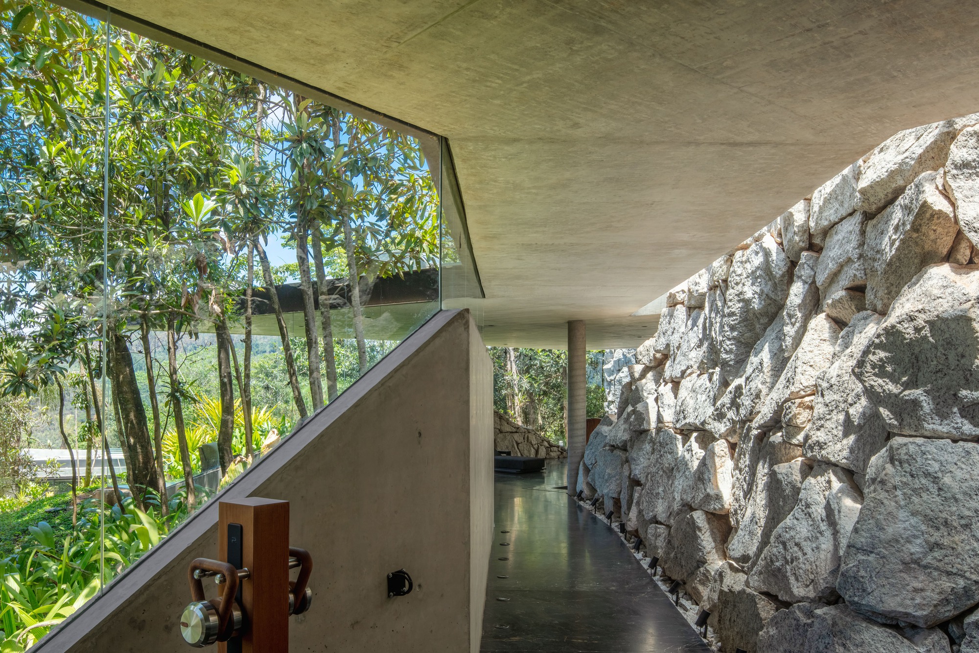 Covered concrete walkway between a glass wall facing treetops and a rough boulder retaining wall, with a slim round column ahead