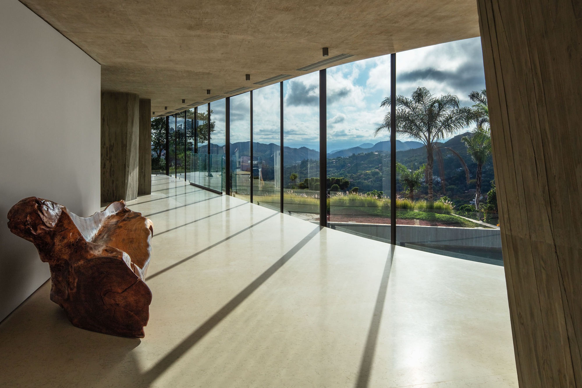 Gallery hall with floor-to-ceiling glazing framing distant mountains and a palm tree, with a carved root-wood seat in the foreground