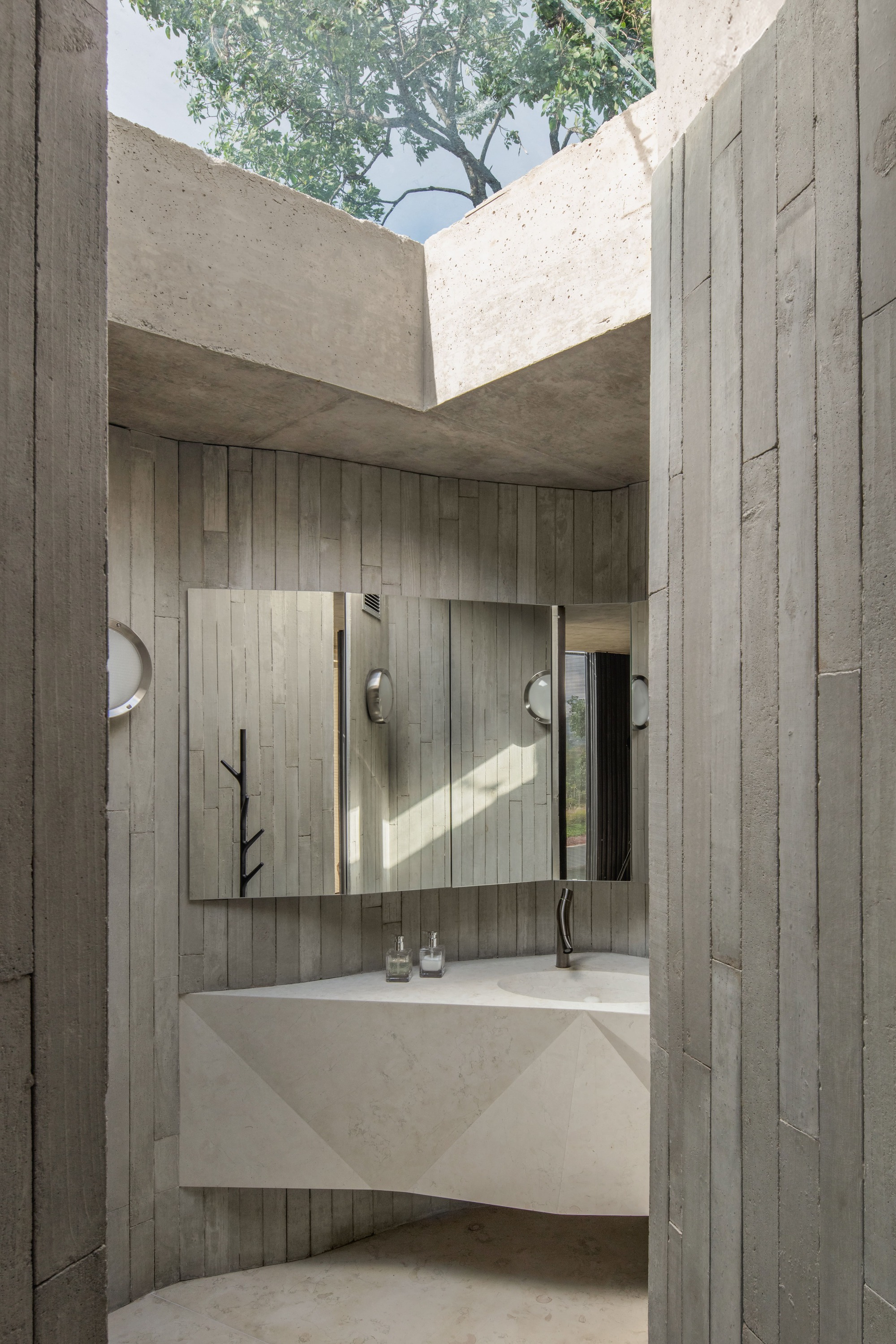 Board-formed concrete washroom with a faceted stone basin, mirror panel and an open skylight showing tree branches overhead