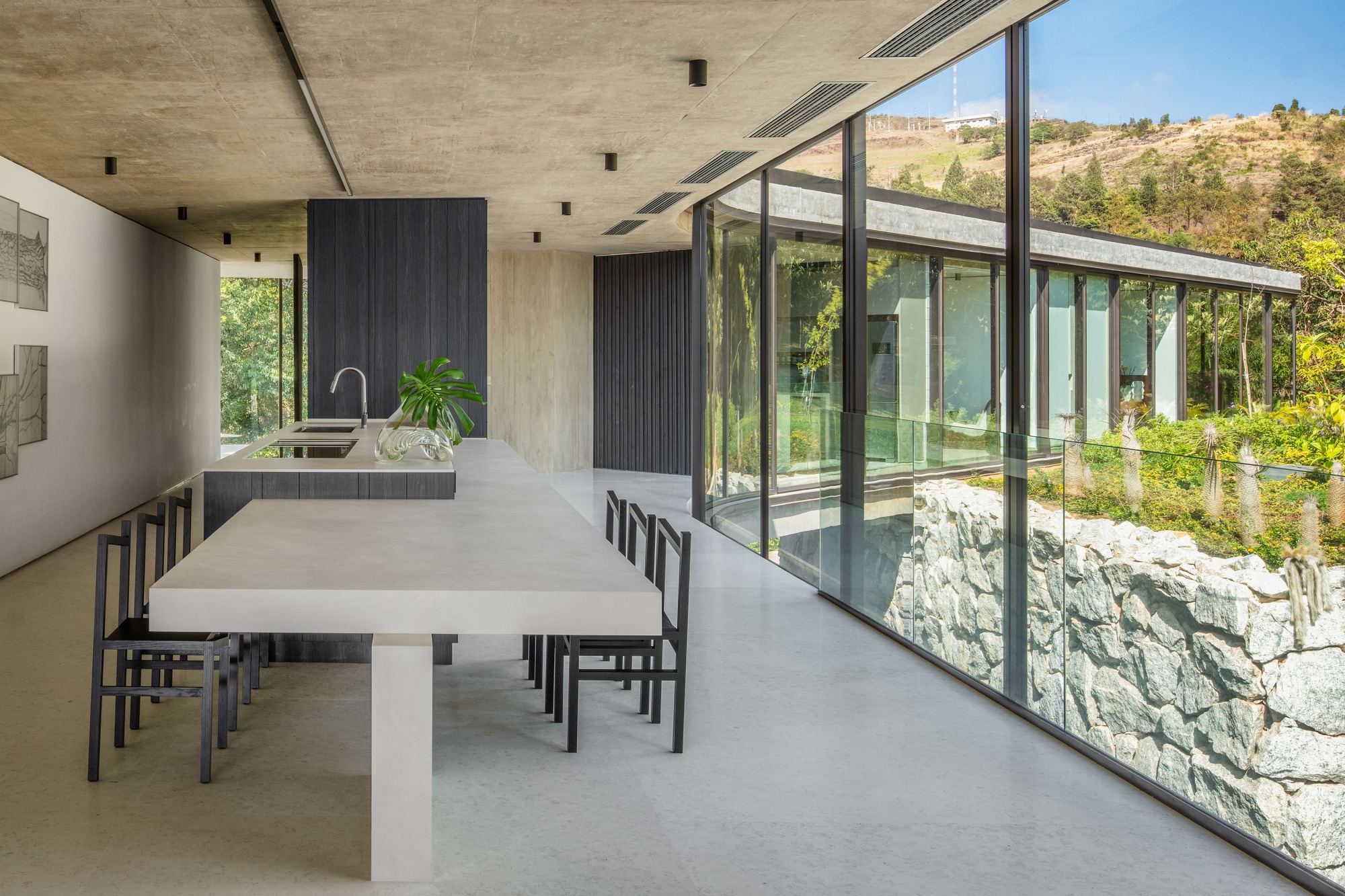 Open kitchen and long stone dining table beside a glass wall facing a rough stone retaining wall and green hillside