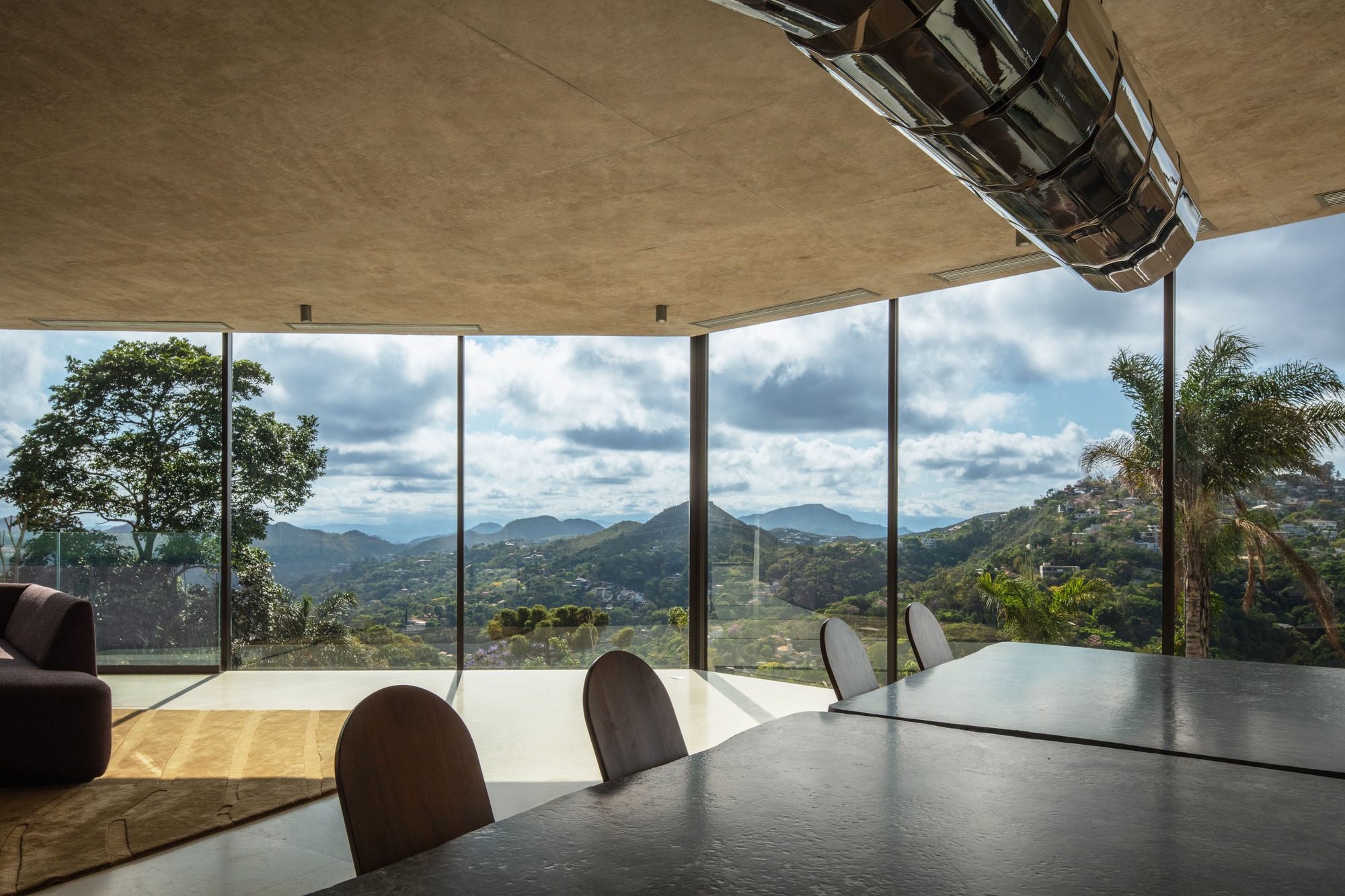 Dining table and chairs under a concrete ceiling, with corner glazing framing distant mountains, a palm and scattered clouds