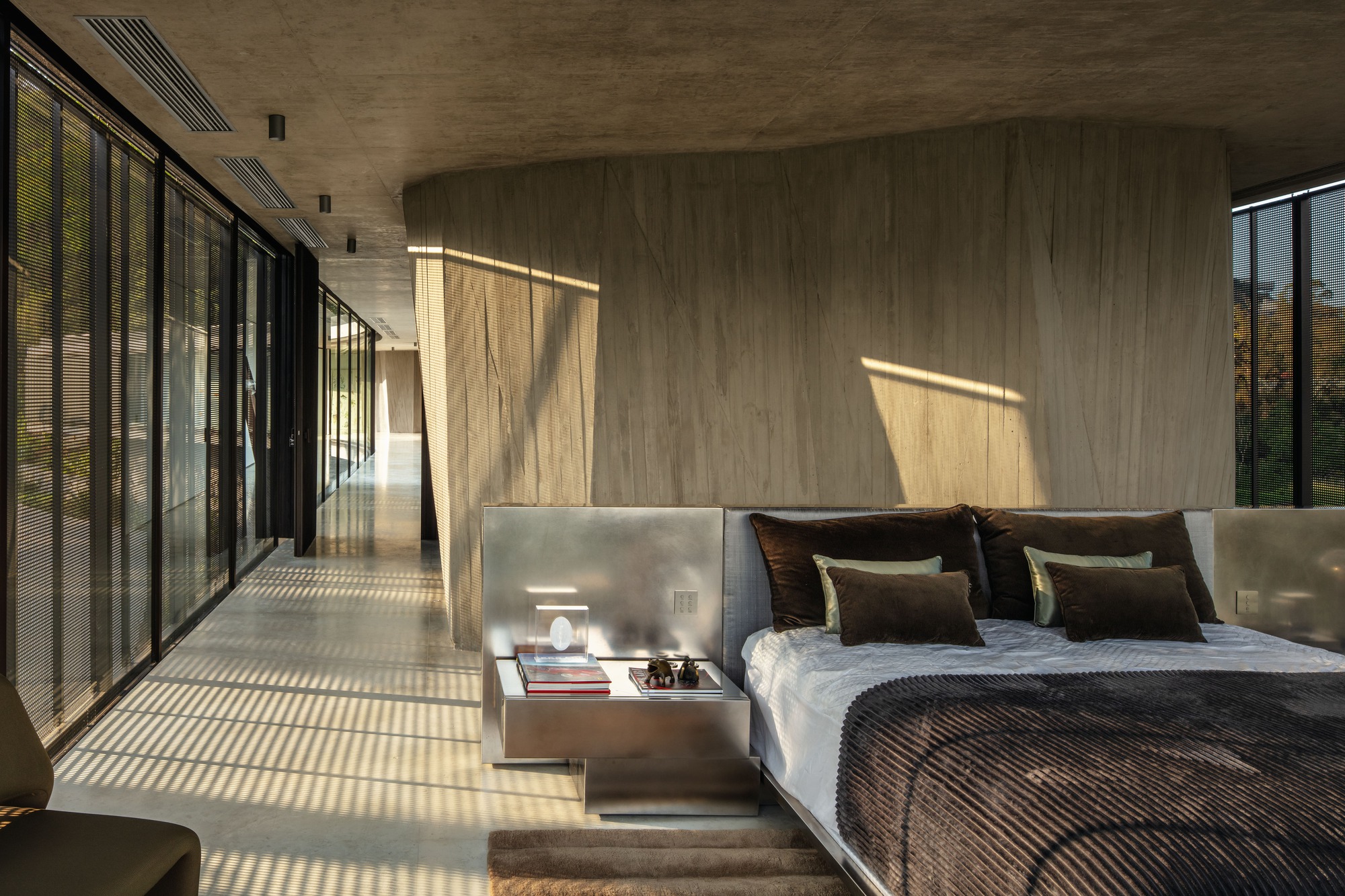 Bedroom with a steel-clad bedhead and nightstand against a curved timber-marked concrete wall, striped by afternoon sun through blinds