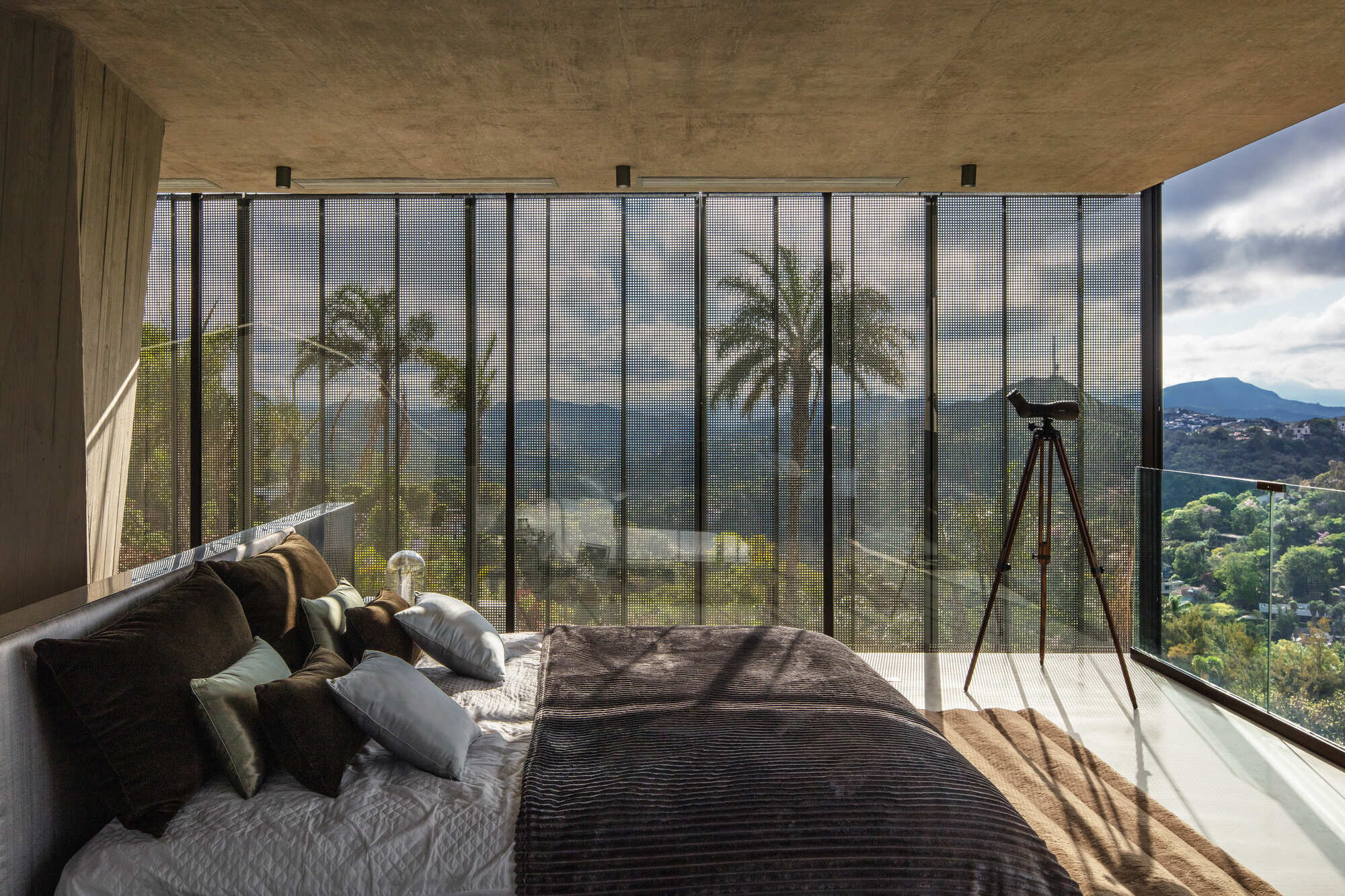 Bed facing a full-height glass wall with a tripod telescope, palms and hazy hills reflected in the panes