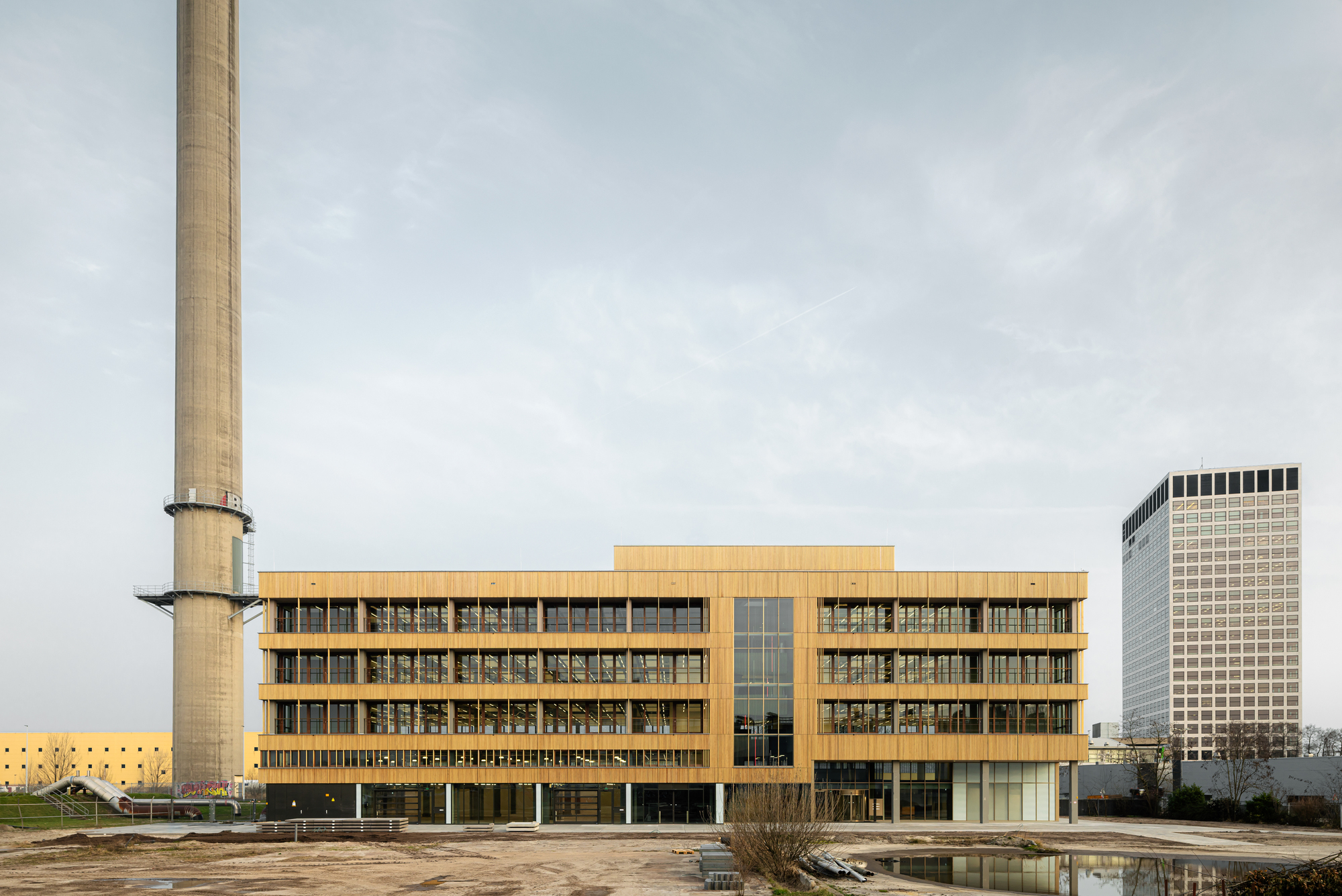Five-storey timber-clad office block beside a tall concrete chimney, with a pond and bare ground in front under an overcast sky