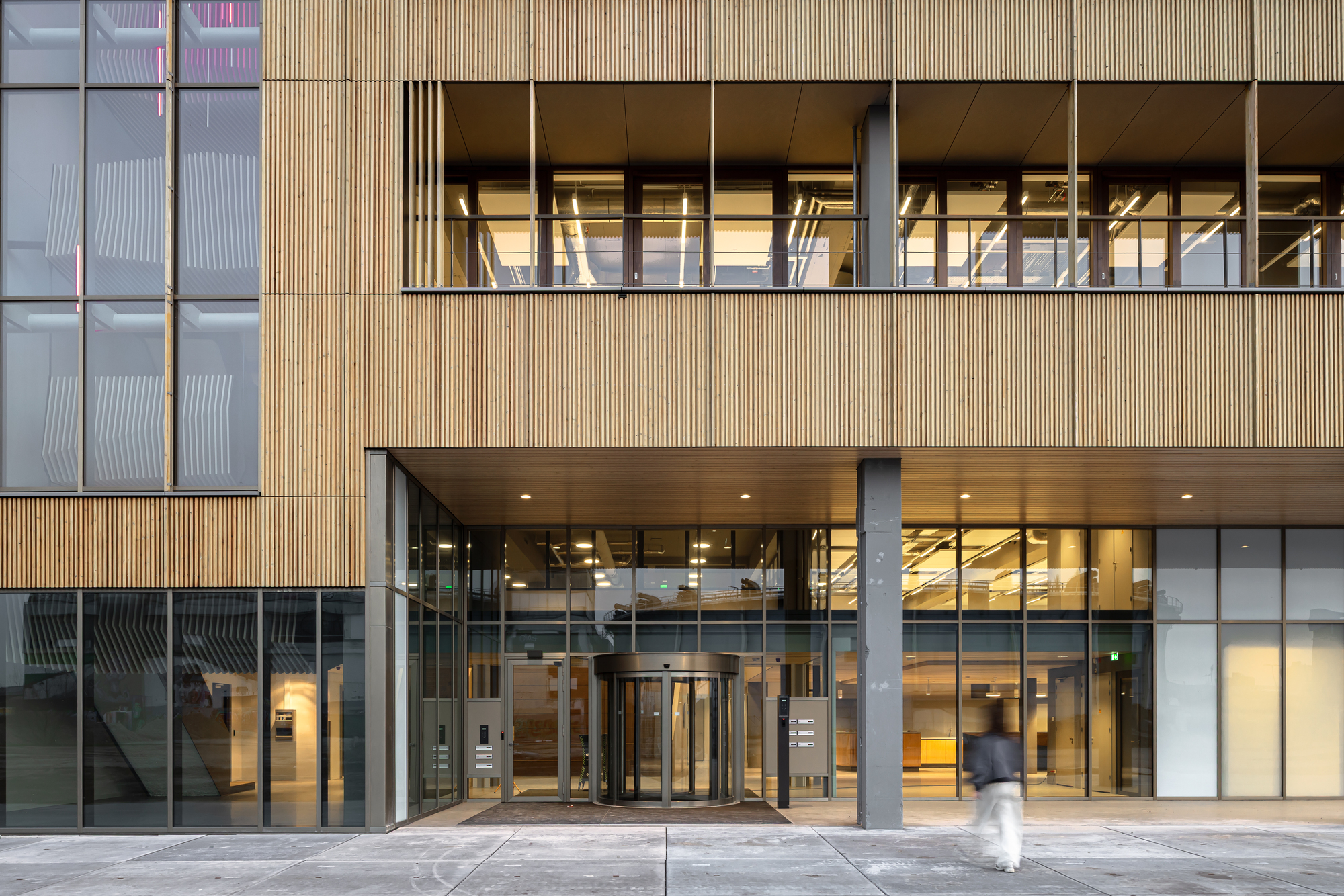 Ground-floor entrance with a revolving door beneath a timber-slatted facade, a blurred pedestrian crossing the paving in the foreground