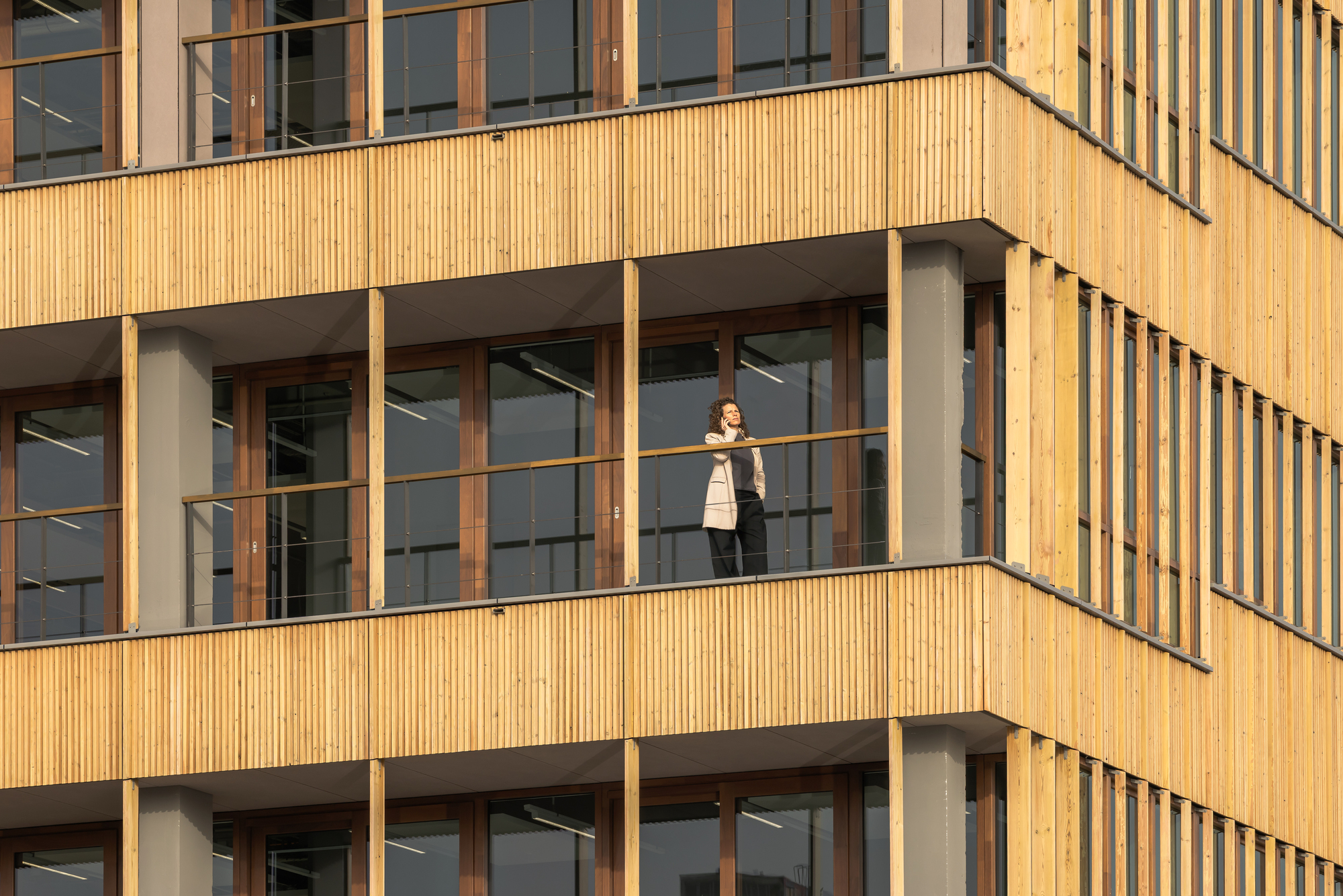 Close view of timber facade bands and grey columns, with a woman on a phone call standing behind the glazed balcony rail in warm light