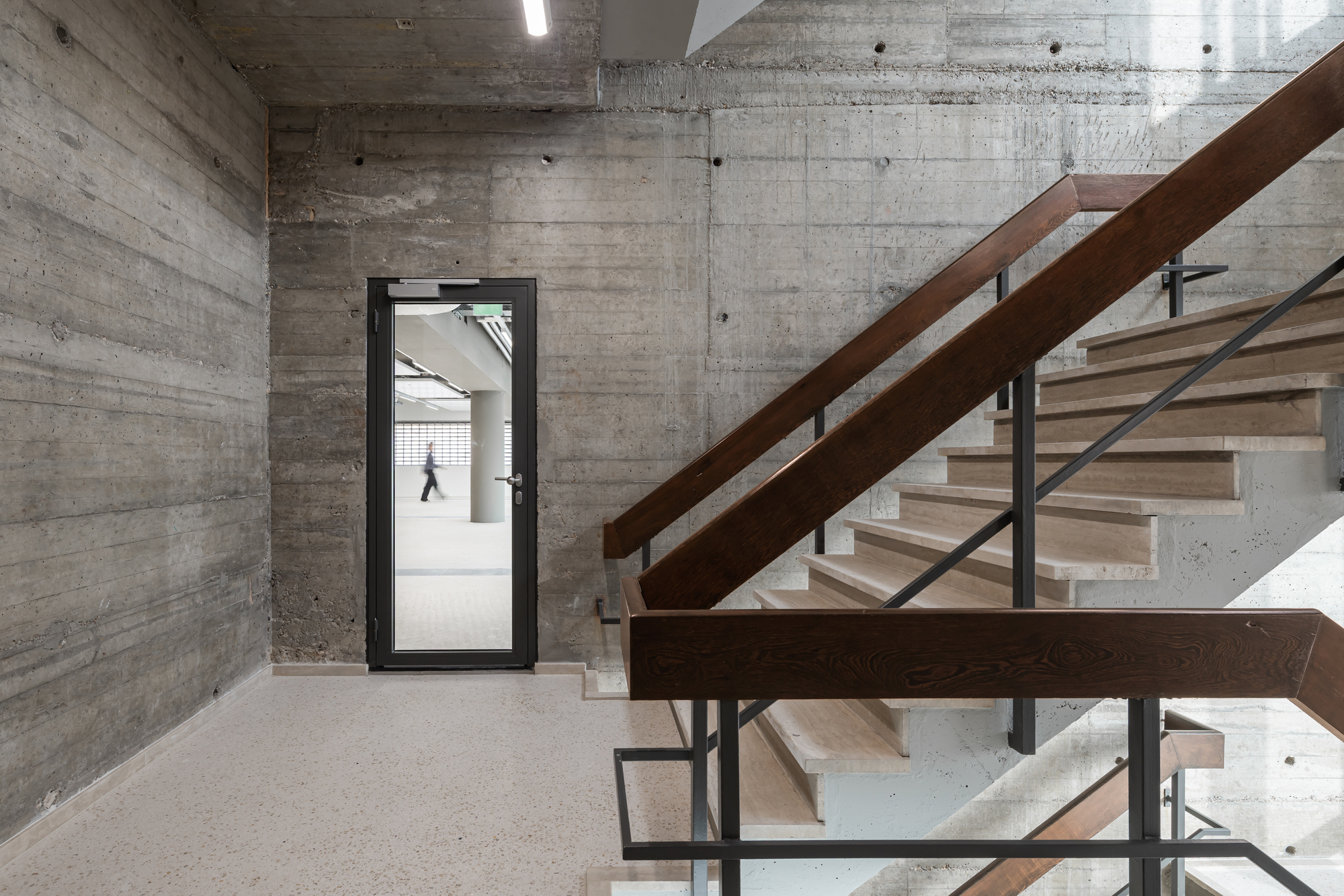 Concrete stairwell with board-marked walls, dark timber handrails and oak treads, a person walking past the glazed door in the background