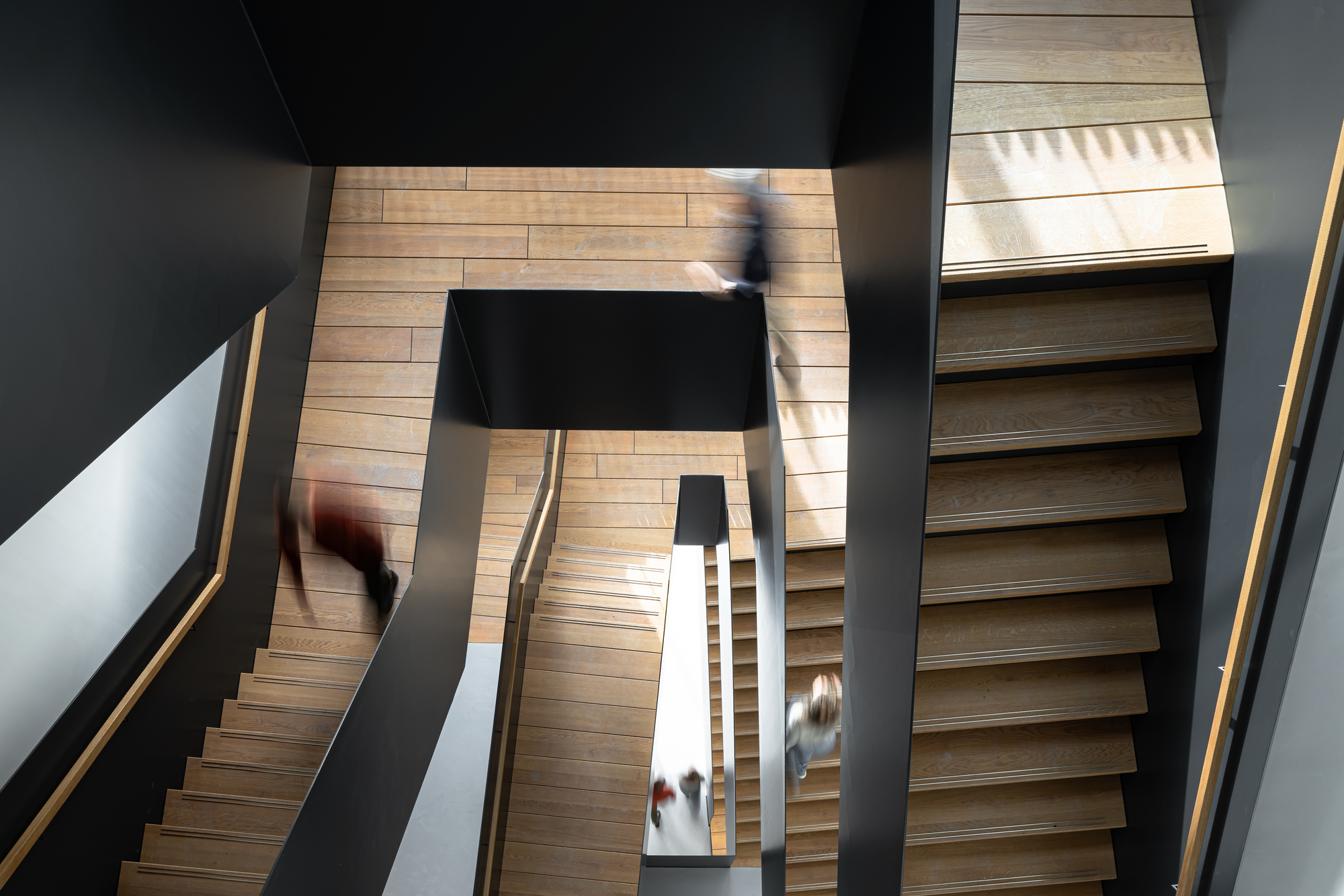 Top-down view of a black-walled stairwell with oak treads and landings, people on the flights blurred in motion, sunlight falling across the upper steps