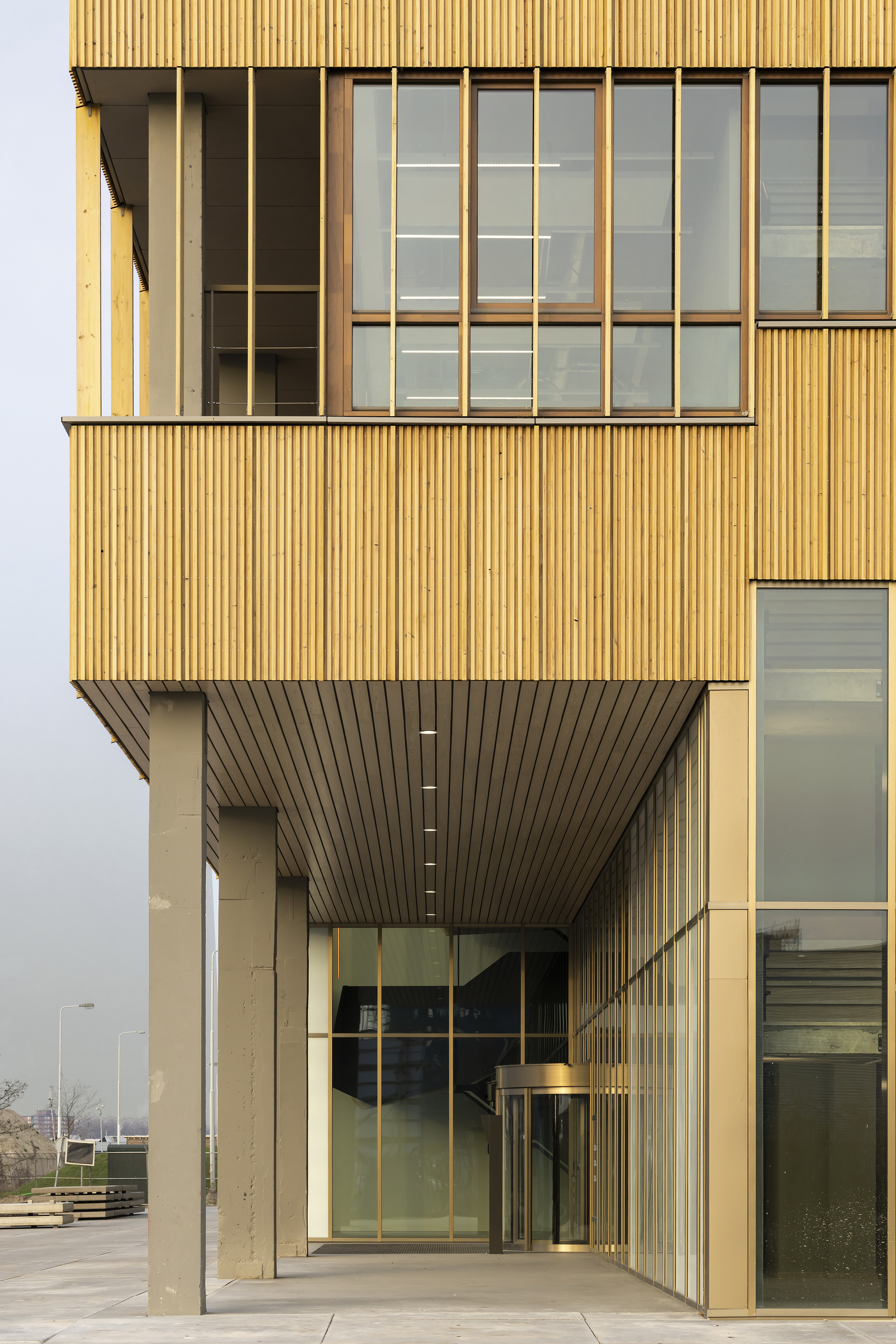 Cantilevered timber-clad upper floors above a recessed glazed entrance with concrete columns, seen under a hazy grey sky