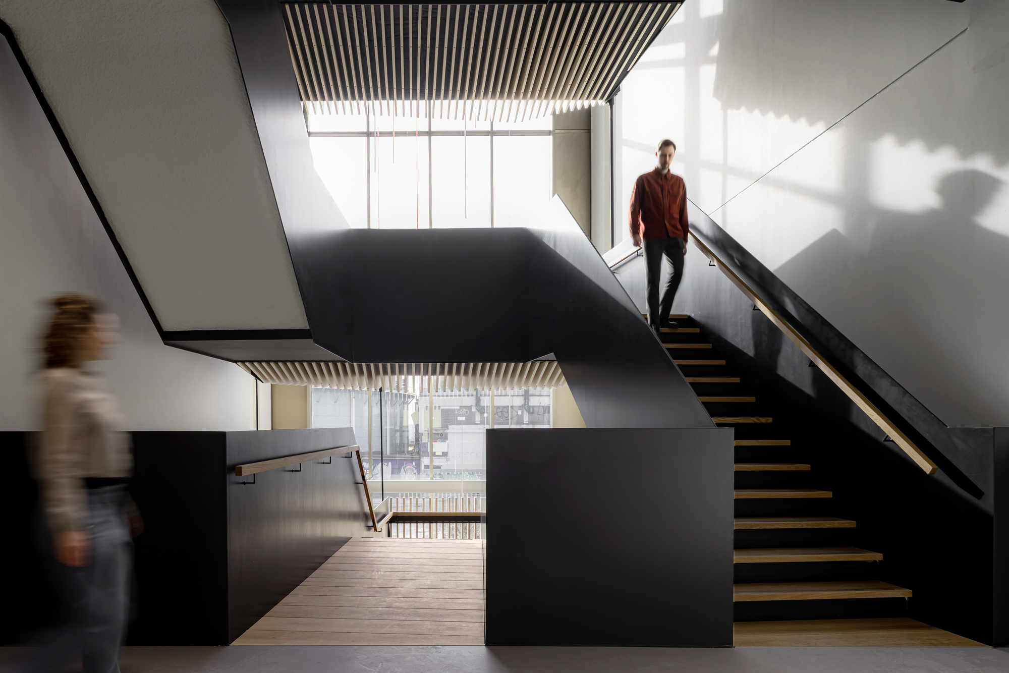 Black-walled stair with timber treads and handrail, a man descending while a blurred figure walks past in daylight