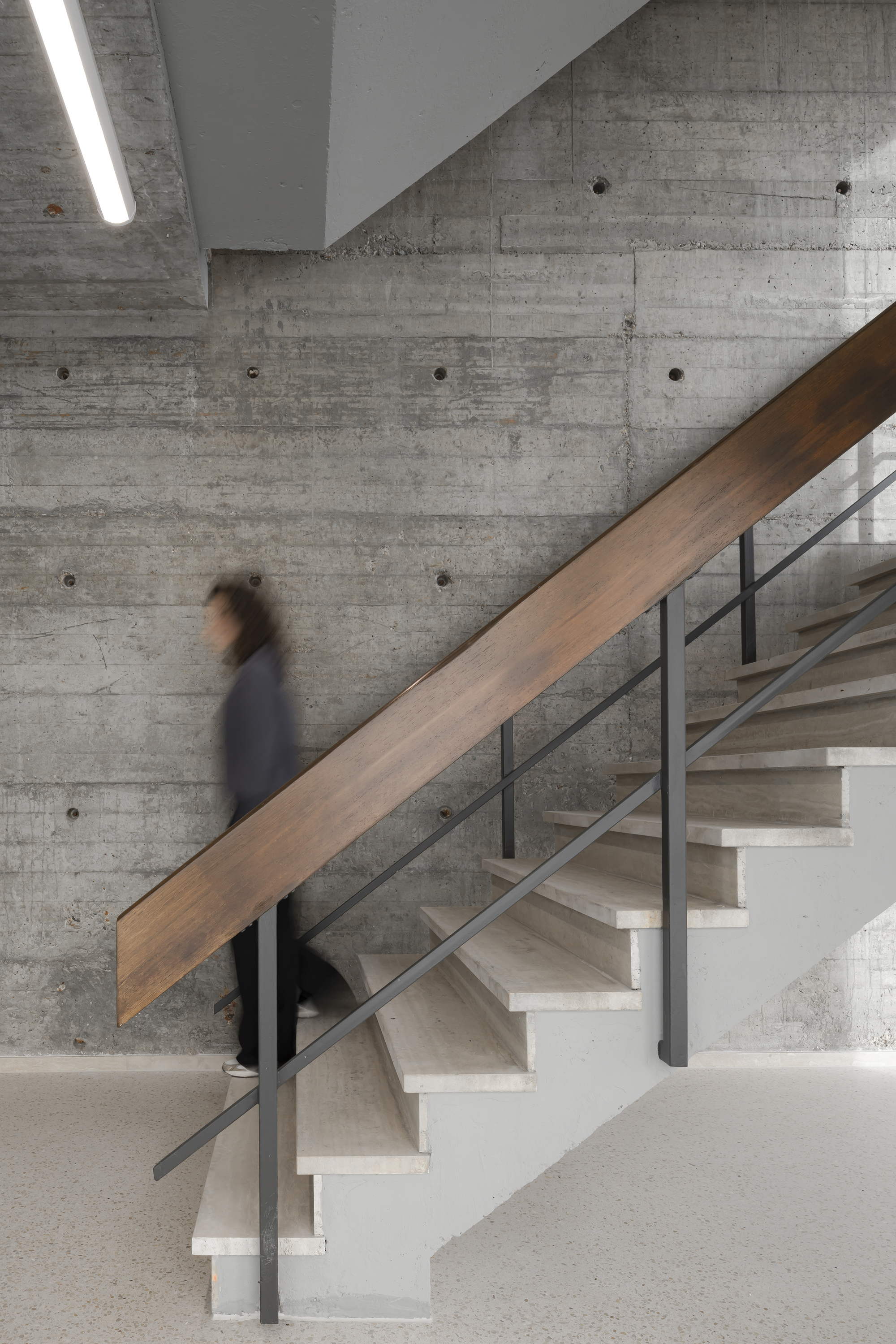 Concrete stair with a dark timber balustrade panel and steel rails against a board-marked wall, a blurred person descending