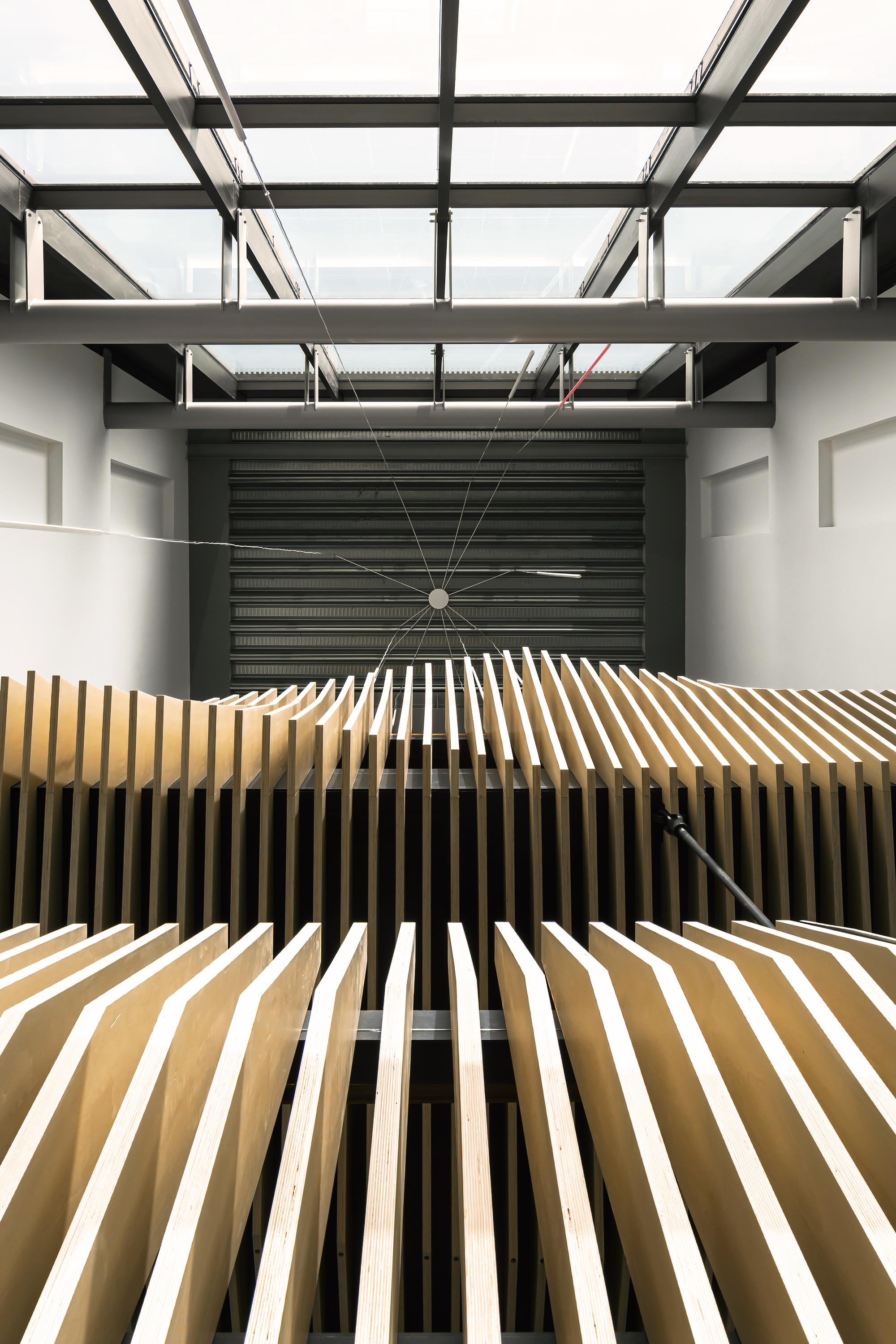 Looking up through a steel-framed skylight at rows of tilted plywood fins above a dark corrugated wall, in bright daylight