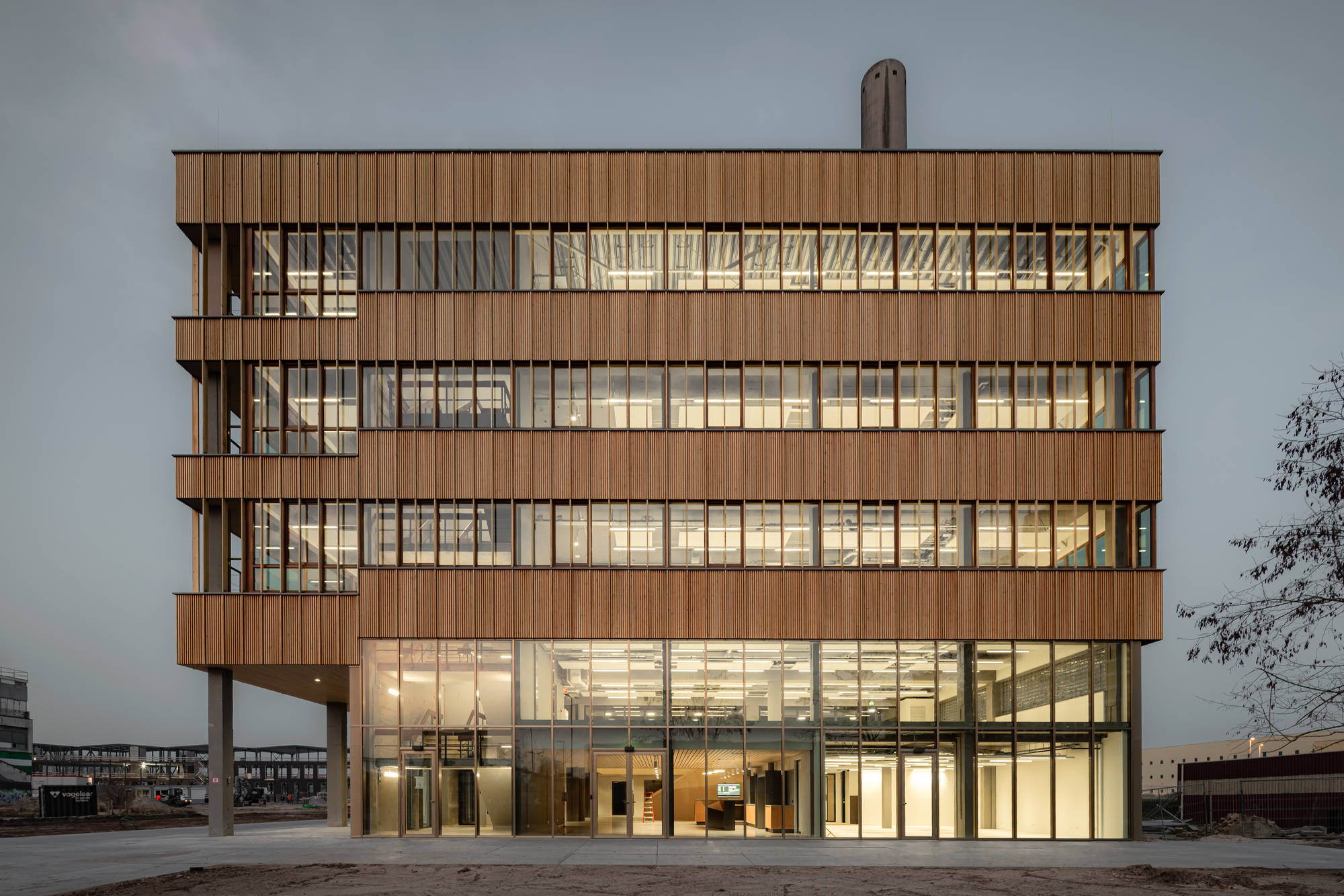Timber-clad five-storey block with a glazed ground floor and a stub chimney behind, lit from within at dusk