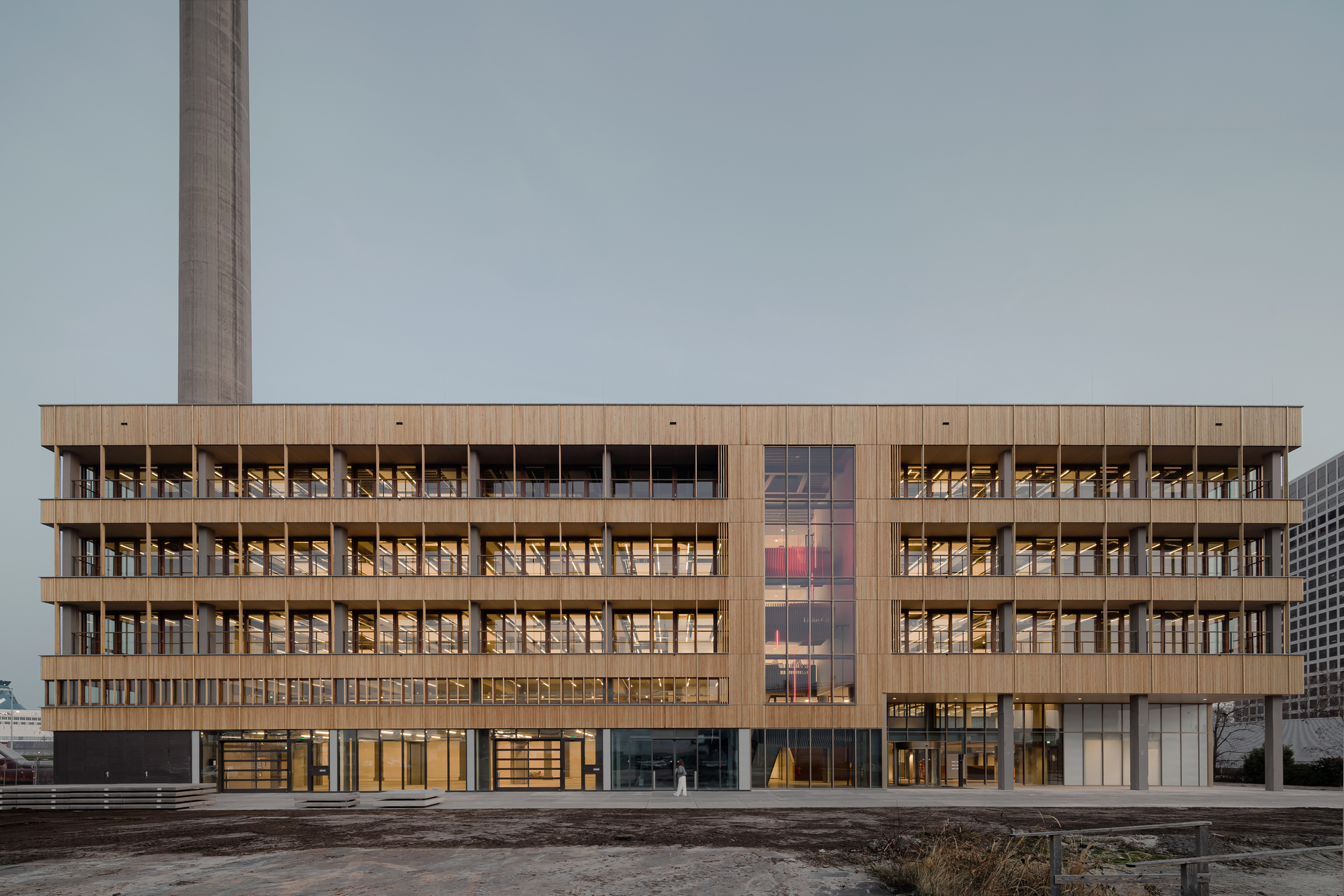 Long timber facade with rows of lit offices, a tall glazed bay and a concrete chimney rising behind, seen at dusk