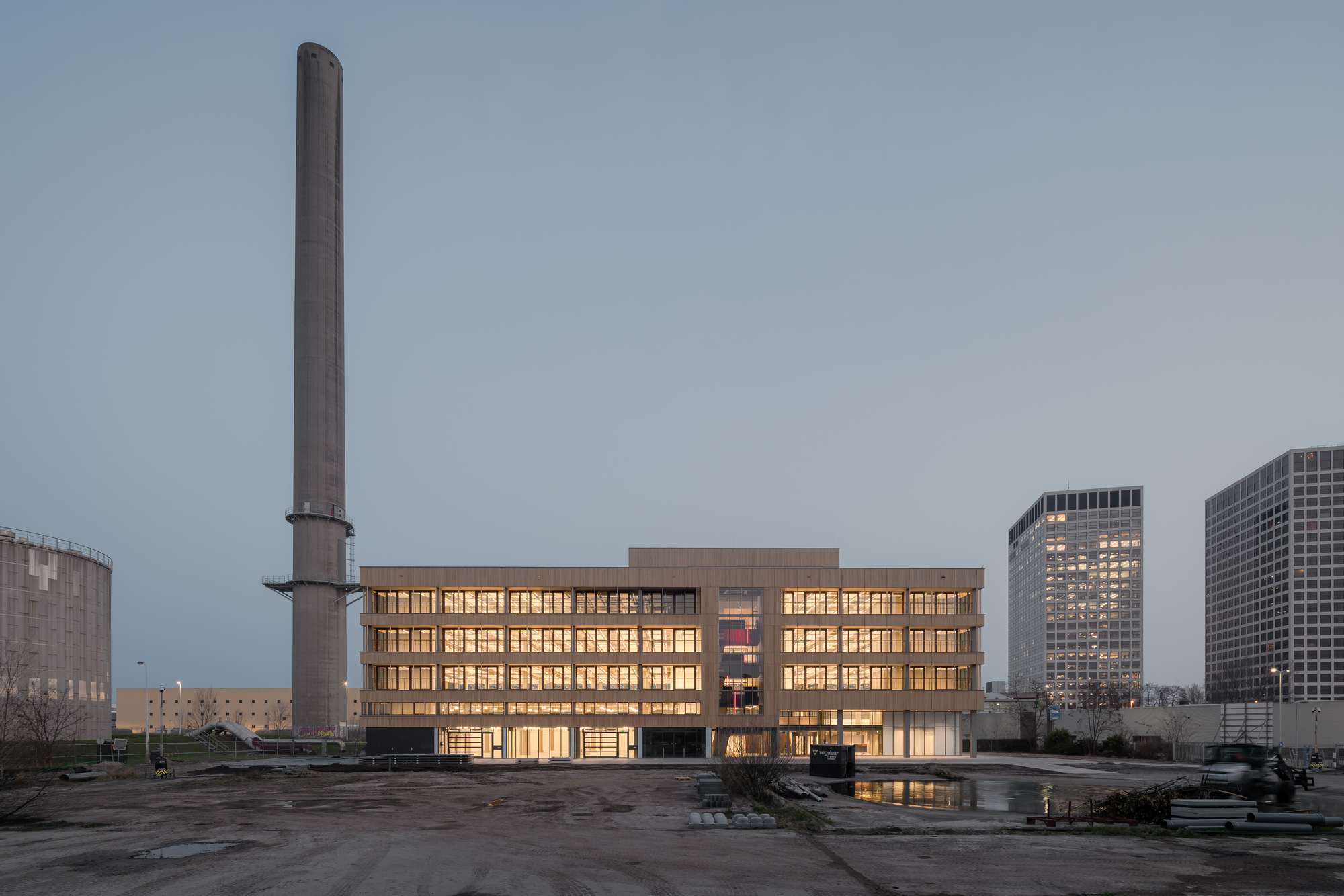 Wide view of the timber-clad block glowing at dusk beside a tall concrete chimney, with office towers and a puddled lot nearby